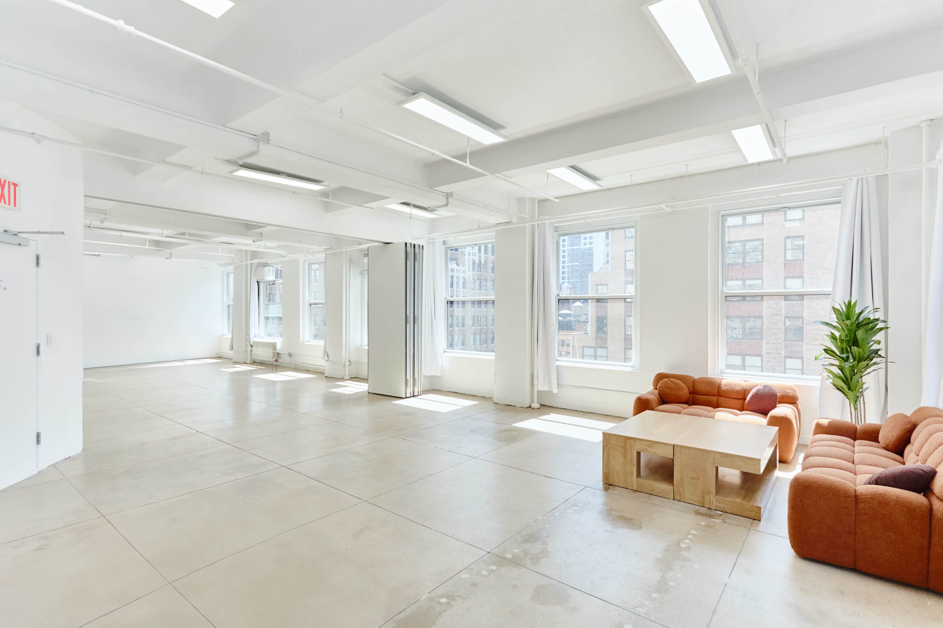 The image shows a spacious, empty interior of a modern office with large windows, concrete flooring, and a seating area featuring a brown couch and a wooden coffee table.