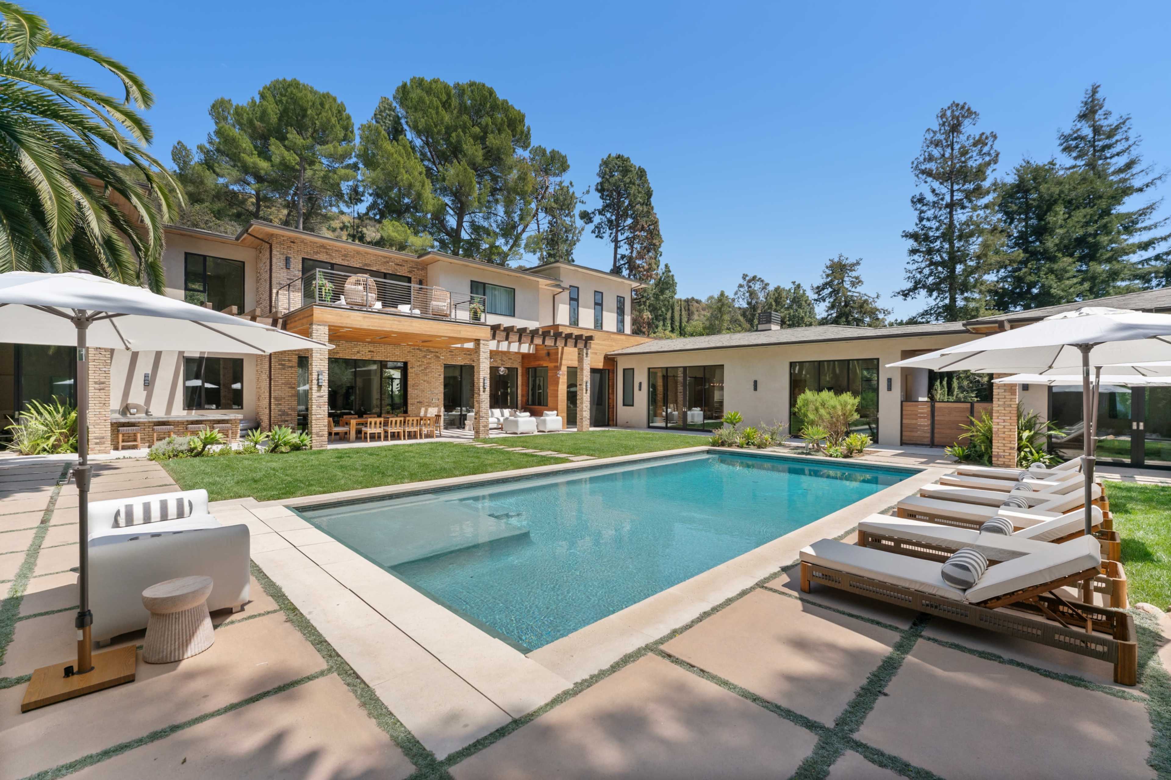 The image features a modern house with a spacious pool area surrounded by lounge chairs and landscaped greenery.