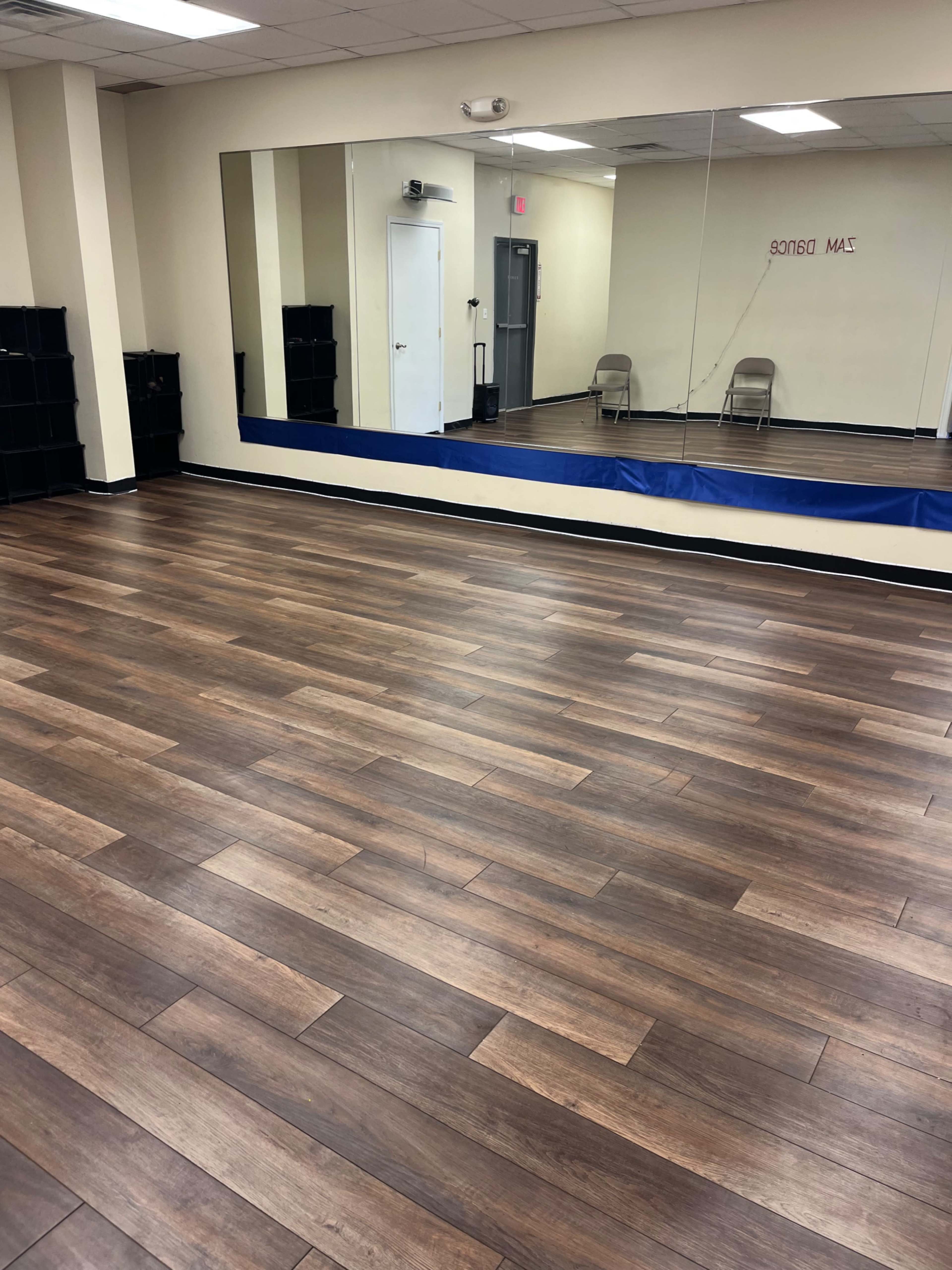 The image shows an empty dance studio with wooden floors, a large mirror on one wall, and two folding chairs in the background.