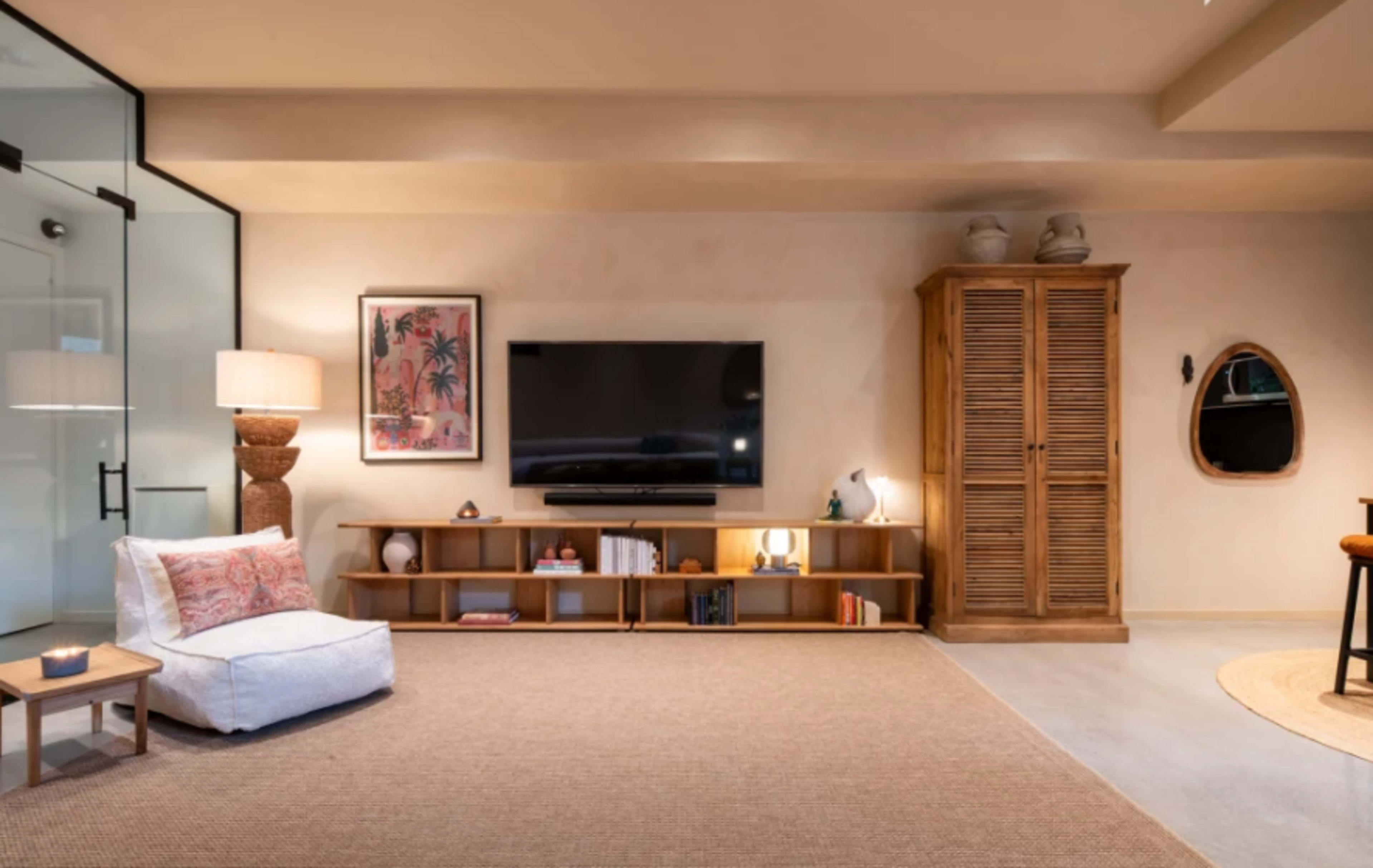 The room features a modern design with a television mounted on the wall, a light-colored rug, a bookshelf, and a mix of decorative elements including a lamp and a wooden cabinet.