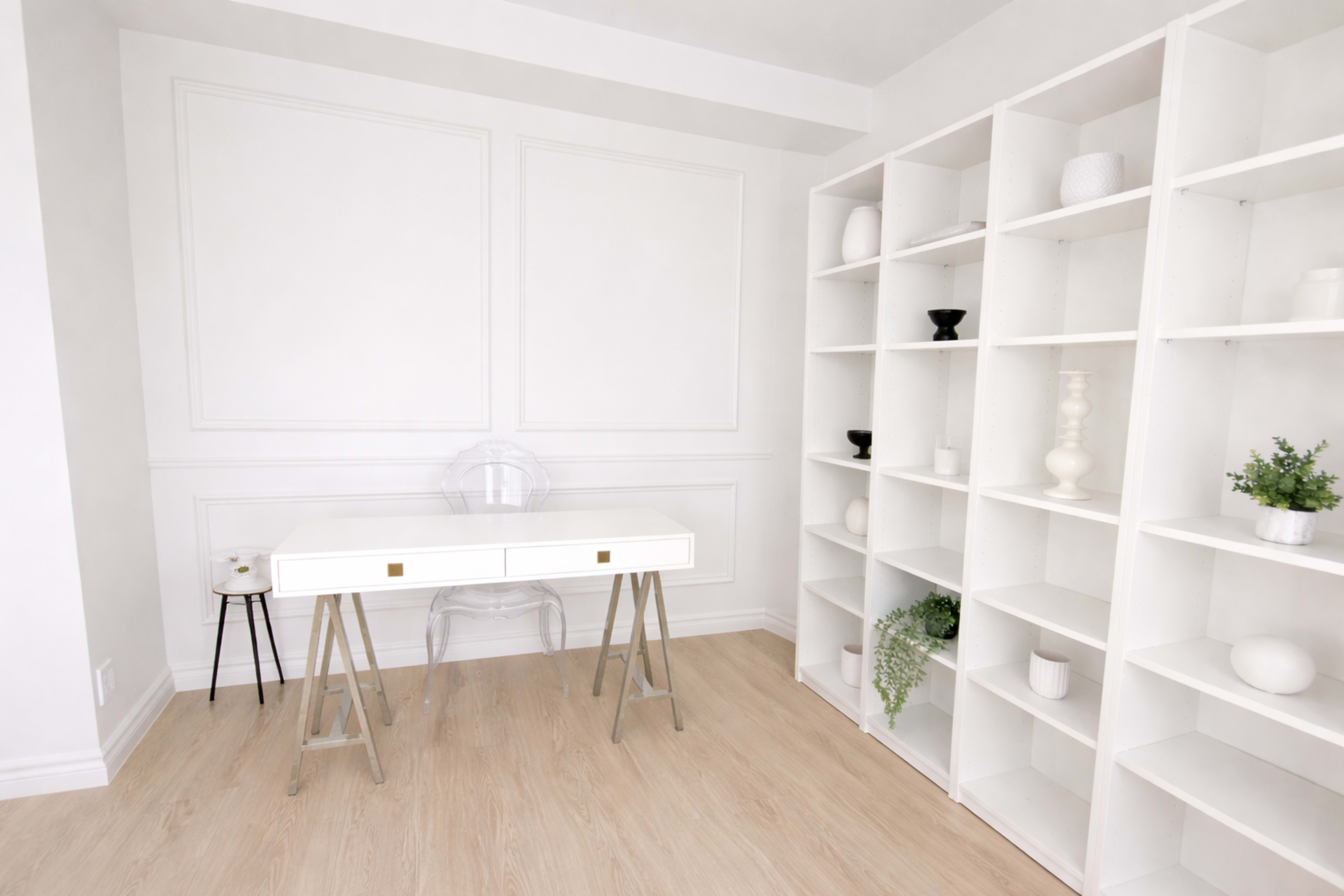The image shows a minimalist office space featuring a white desk with two drawers, a clear chair, and a white shelving unit filled with decorative items and plants.
