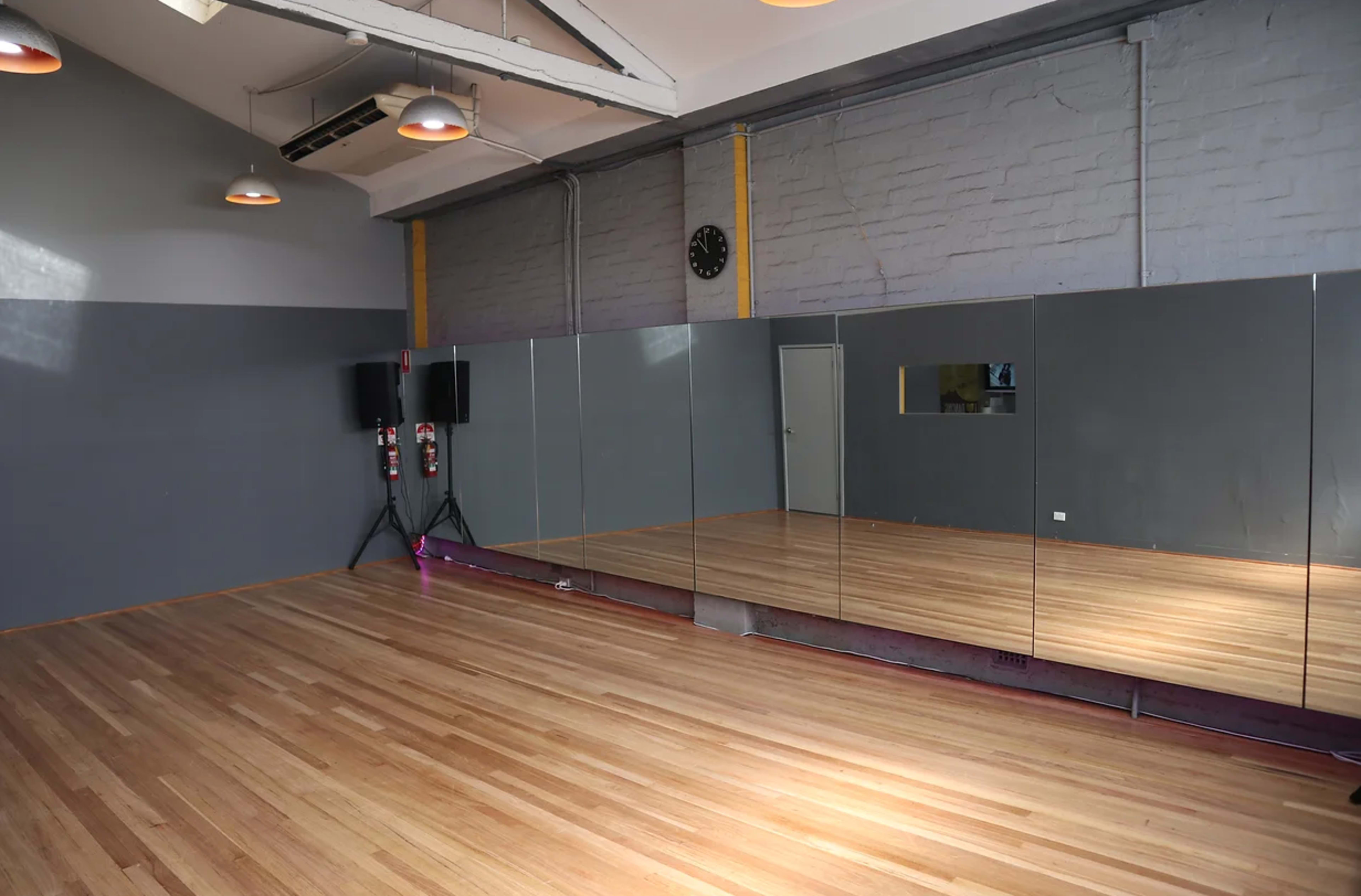 The image shows a spacious dance studio with wooden floors, gray walls, large mirrors, and sound equipment in the corners.