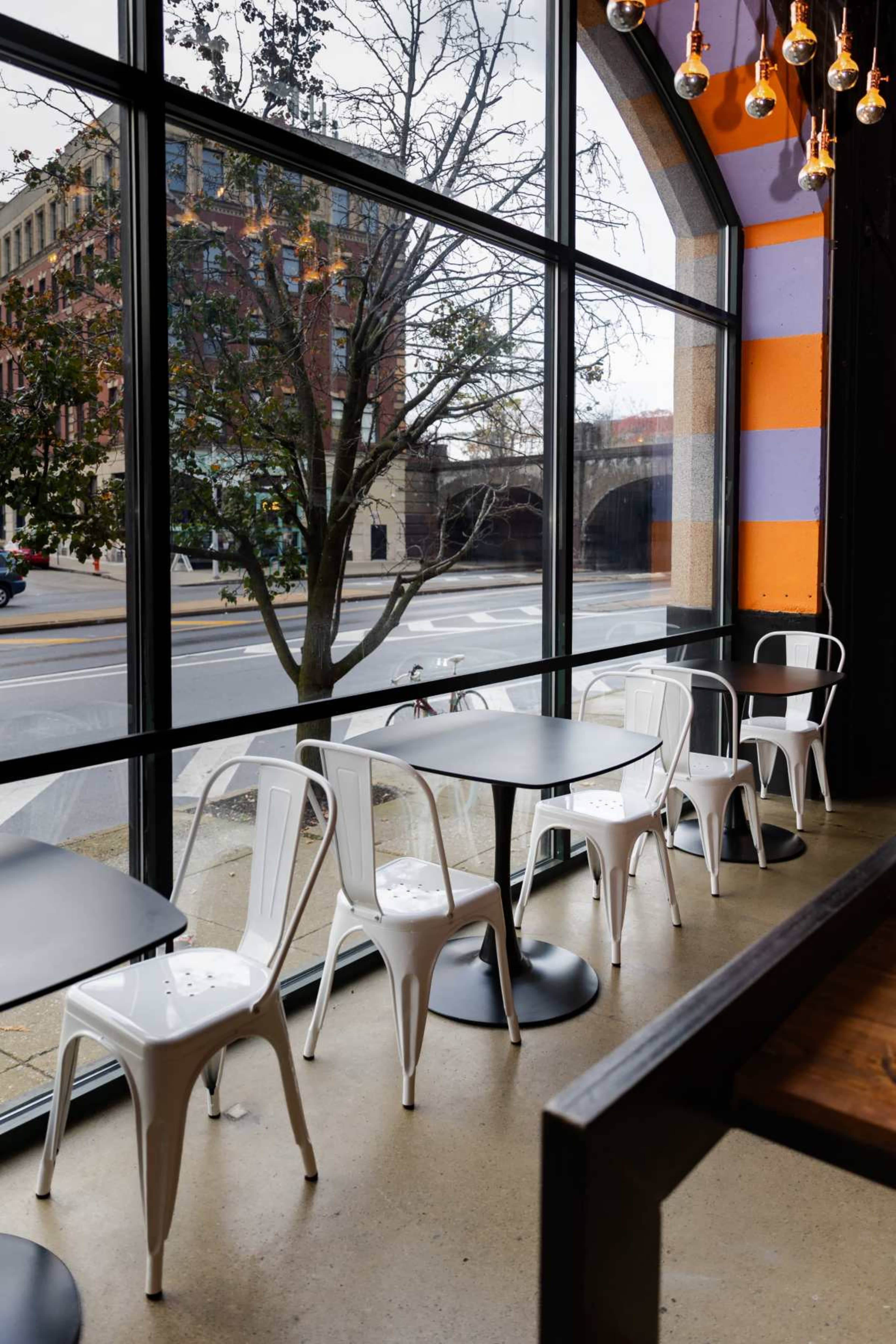 The image shows a modern café interior with white metal chairs and tables arranged near large windows overlooking a street scene.
