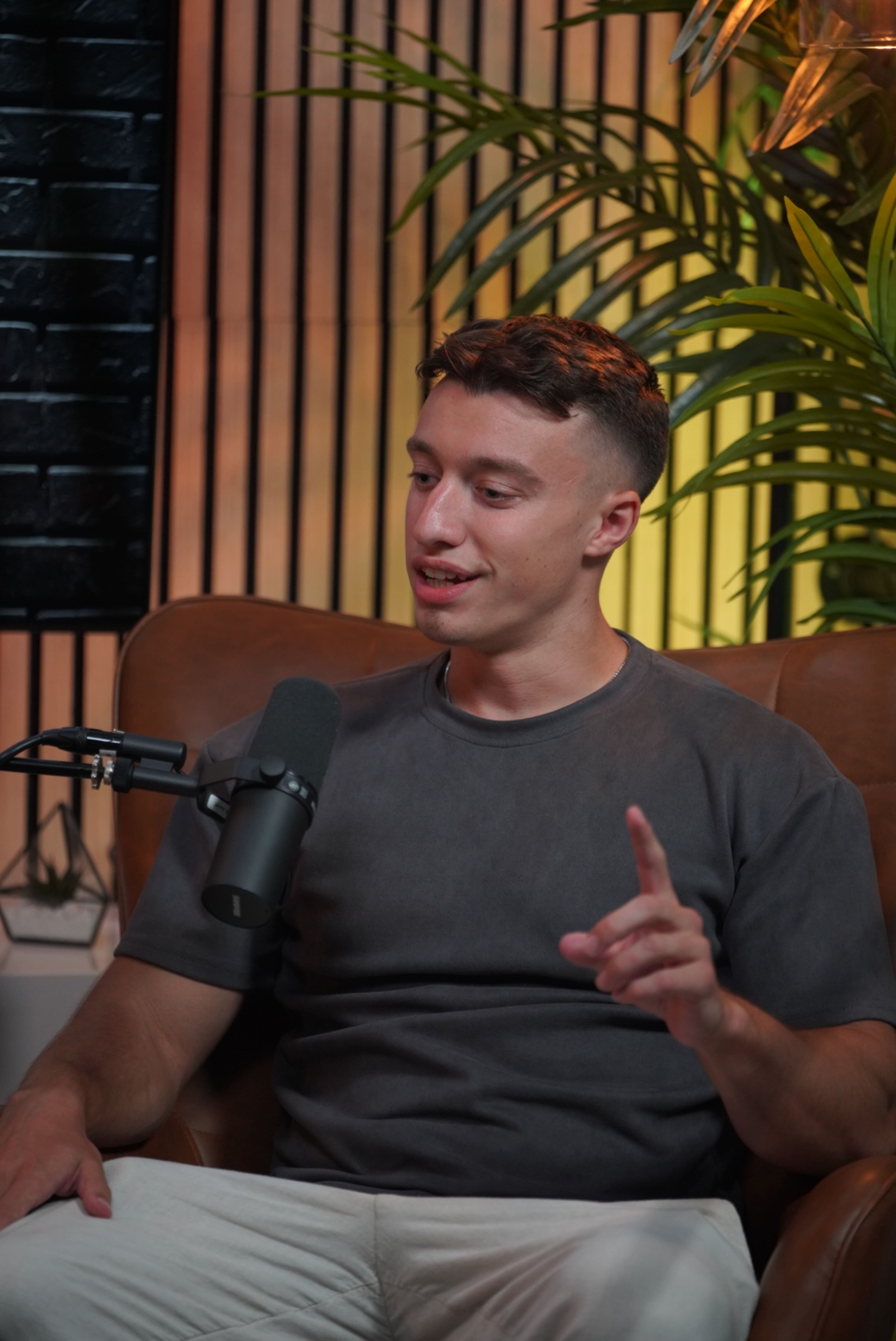 A young man sits in a chair with a microphone in front of him, gesturing while engaged in conversation against a backdrop of plants and a brick wall.