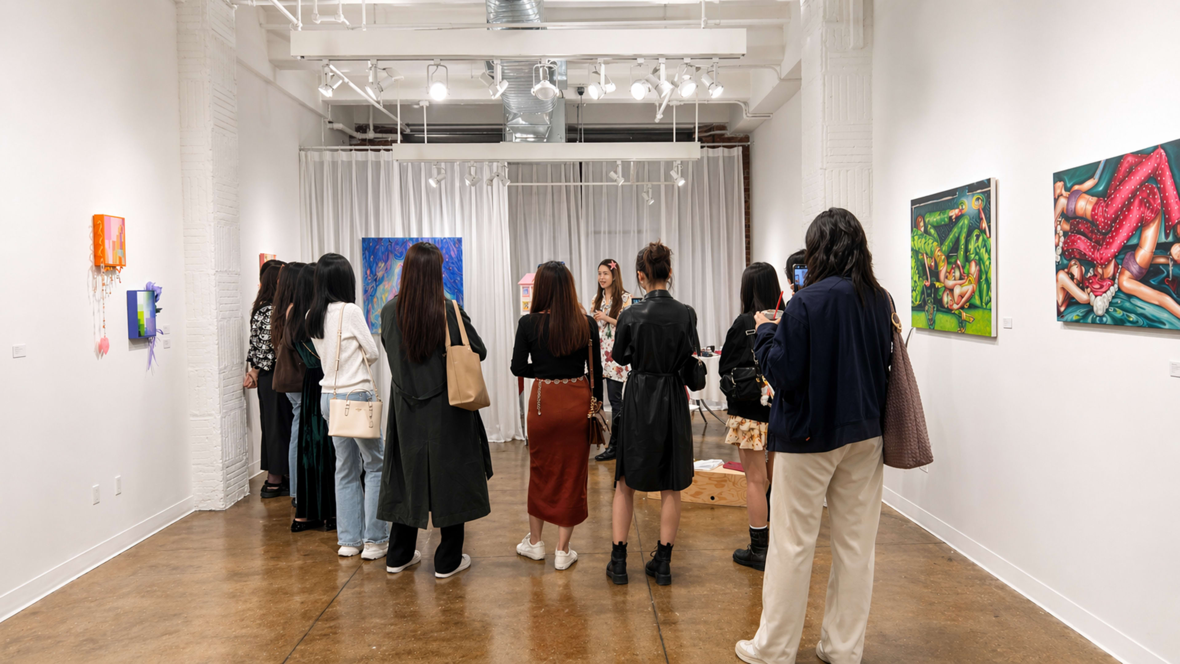 A group of people stands in an art gallery, attentively observing an artist speaking in front of several colorful paintings displayed on the walls.