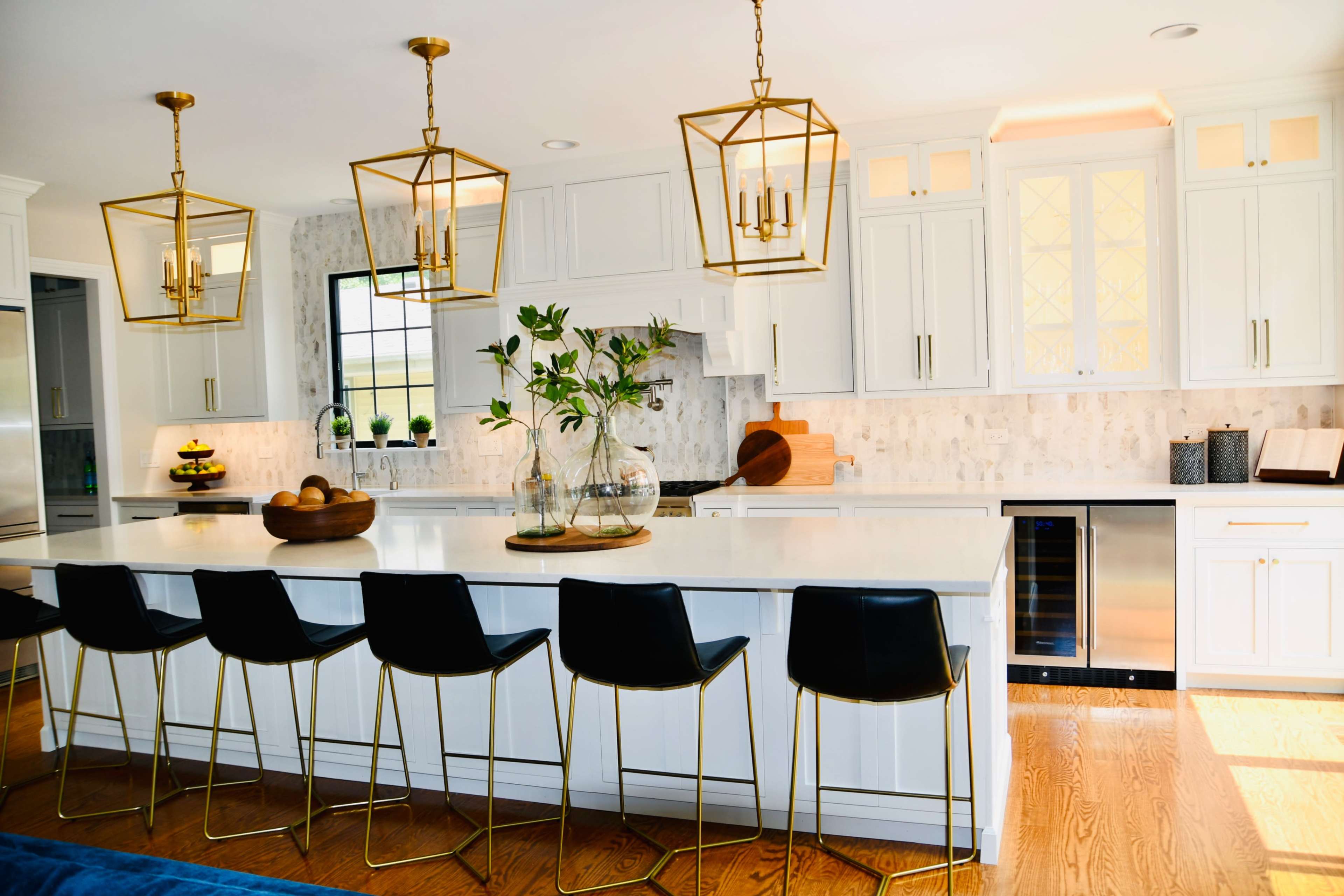 A modern kitchen features a large island with black bar stools, gold pendant lights, and white cabinets, complemented by a light-toned backsplash and stainless steel appliances.