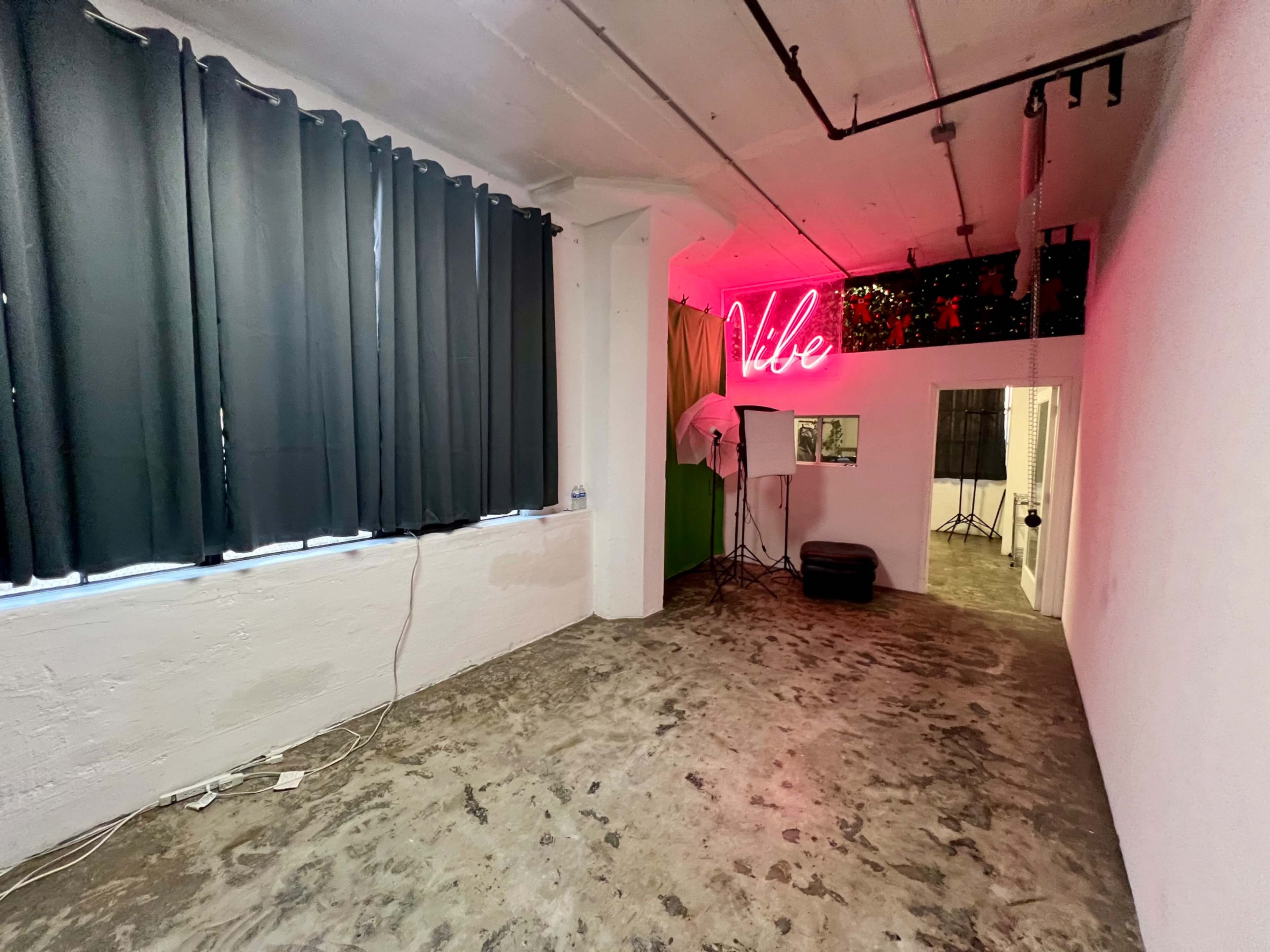 The image shows an empty room with concrete flooring, black curtains, and a neon sign that says "Vibe" on the wall.
