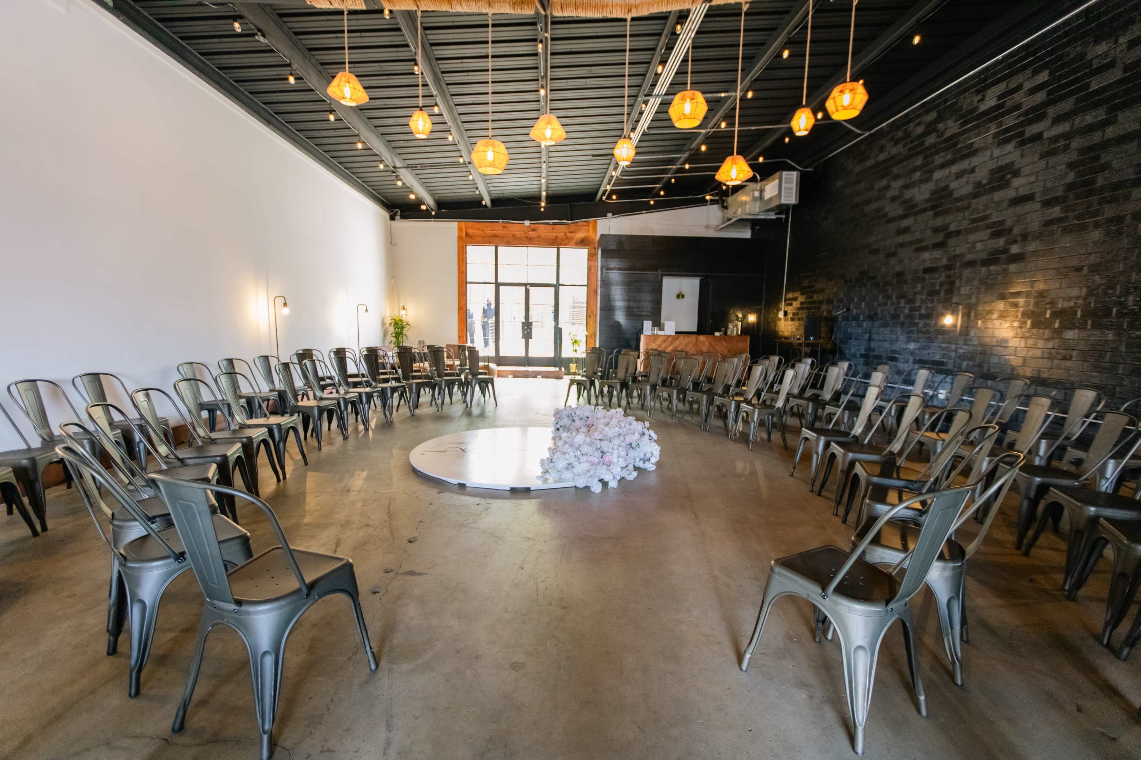 The image shows a spacious venue arranged with metal chairs in a circular formation around a small platform, illuminated by pendant lights.