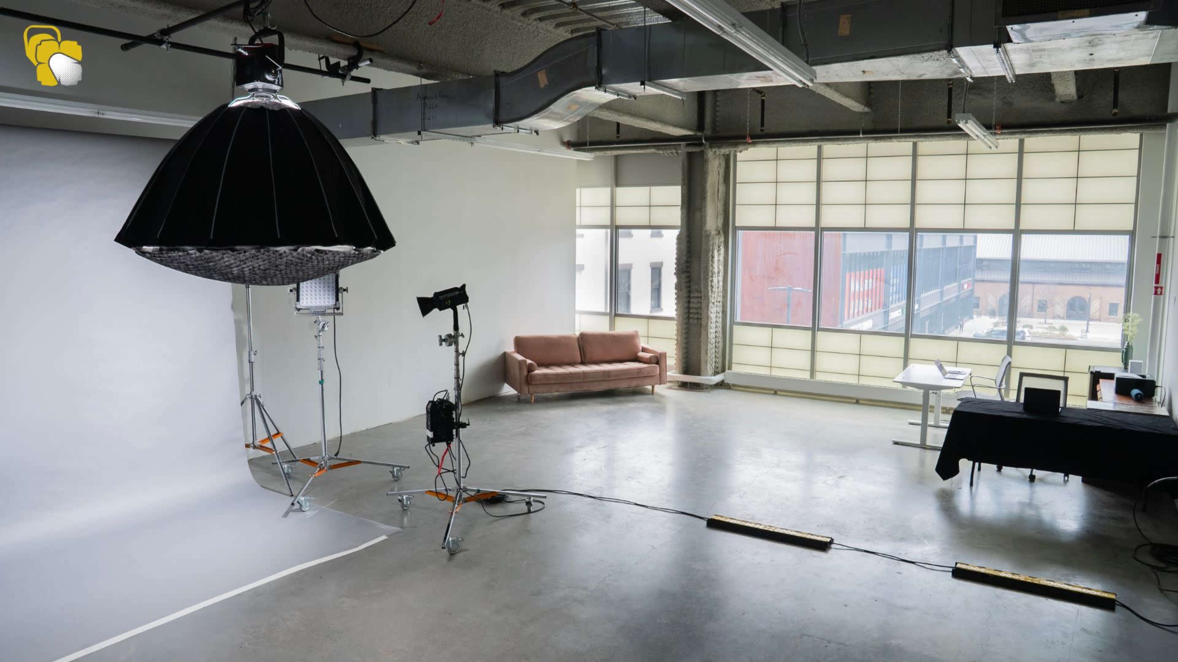 The image shows a spacious photography studio with a large light fixture, a brown couch, and a table set up near a window with natural light.