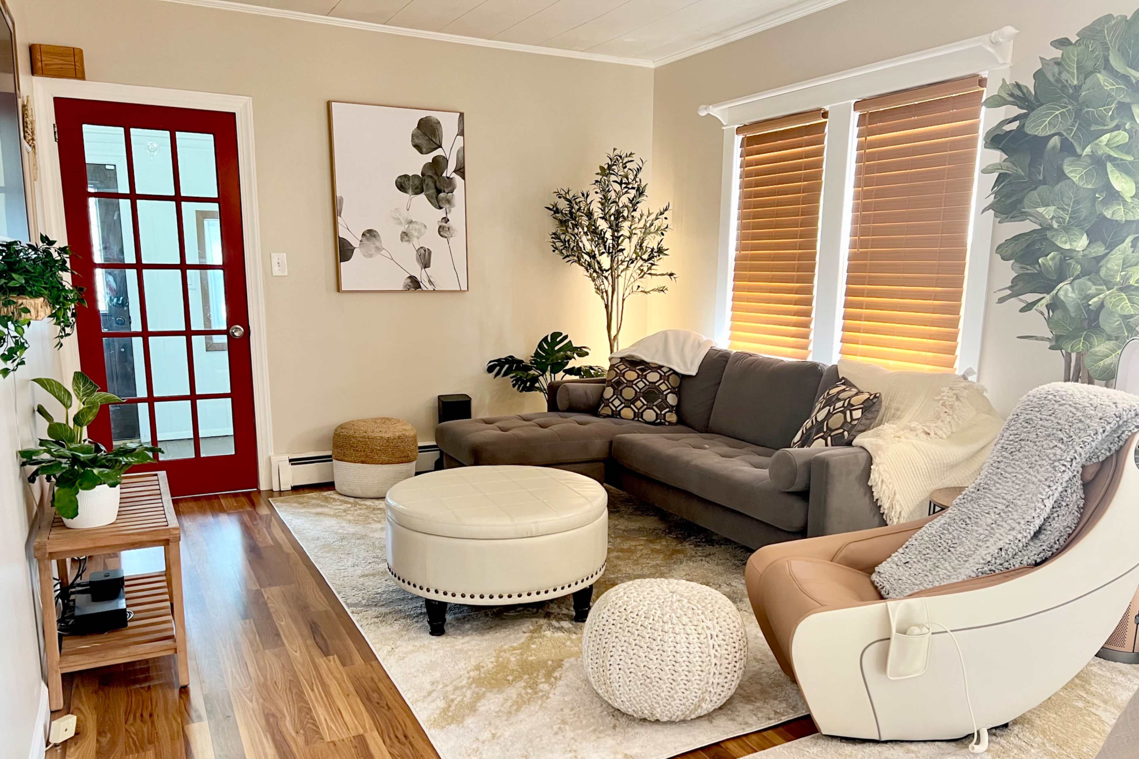 The living room features a gray sectional sofa, a round ottoman, a cozy armchair, and natural light streaming through the window with wooden blinds.