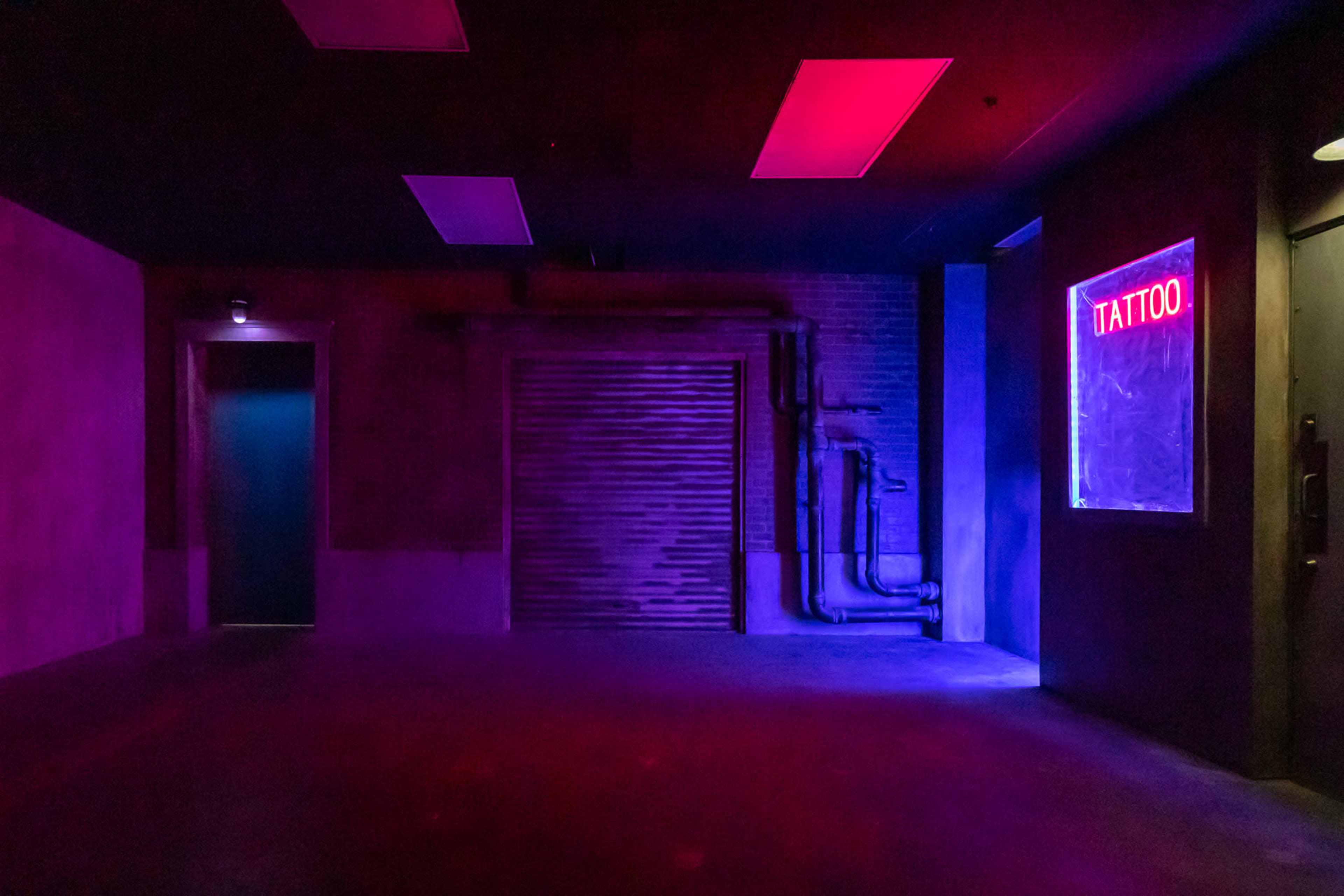 The dimly lit space features a closed garage door, neon-lit "TATTOO" sign, and contrasting colored walls.