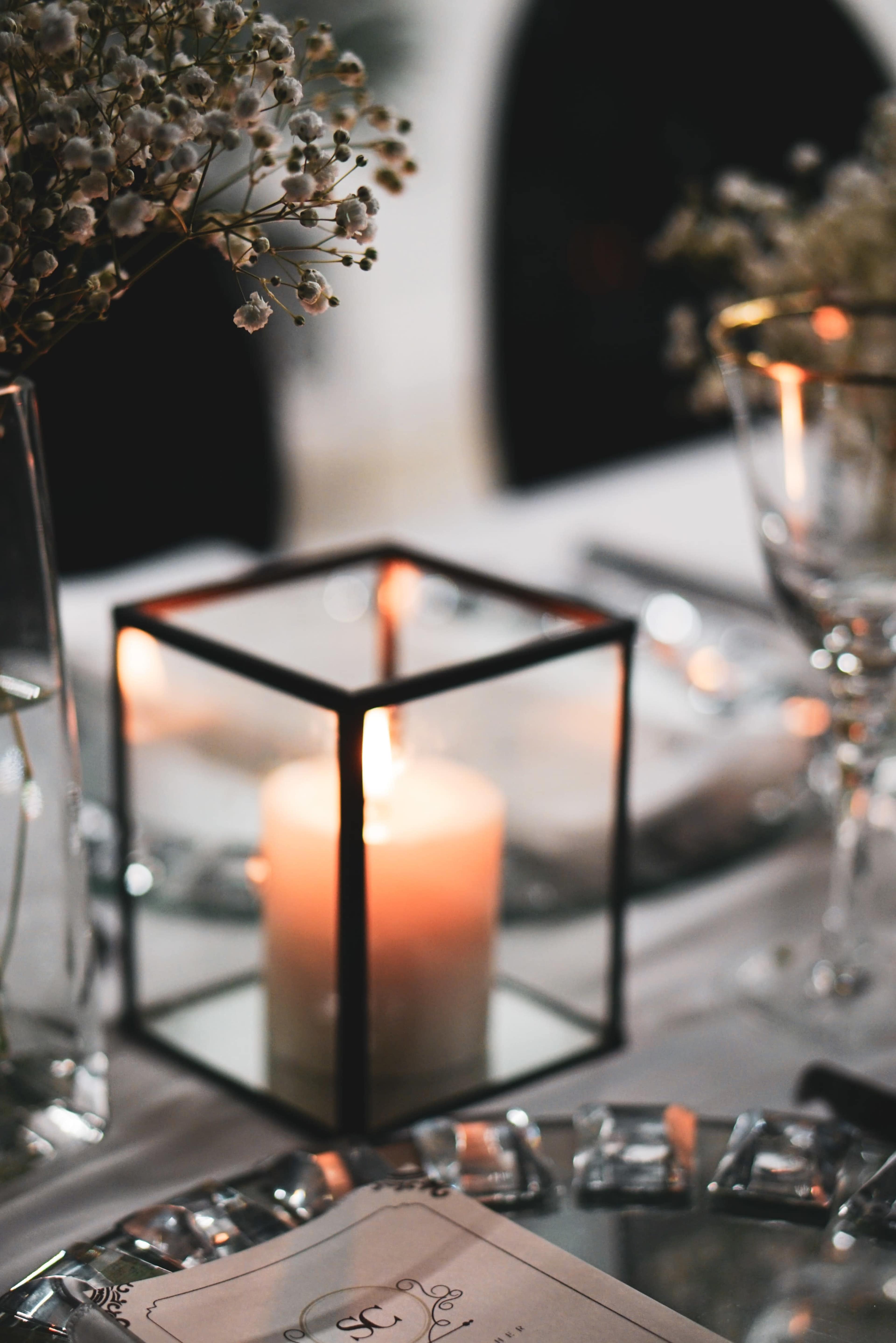 A glass candle holder with a lit candle sits on a table decorated with flowers and dinnerware.