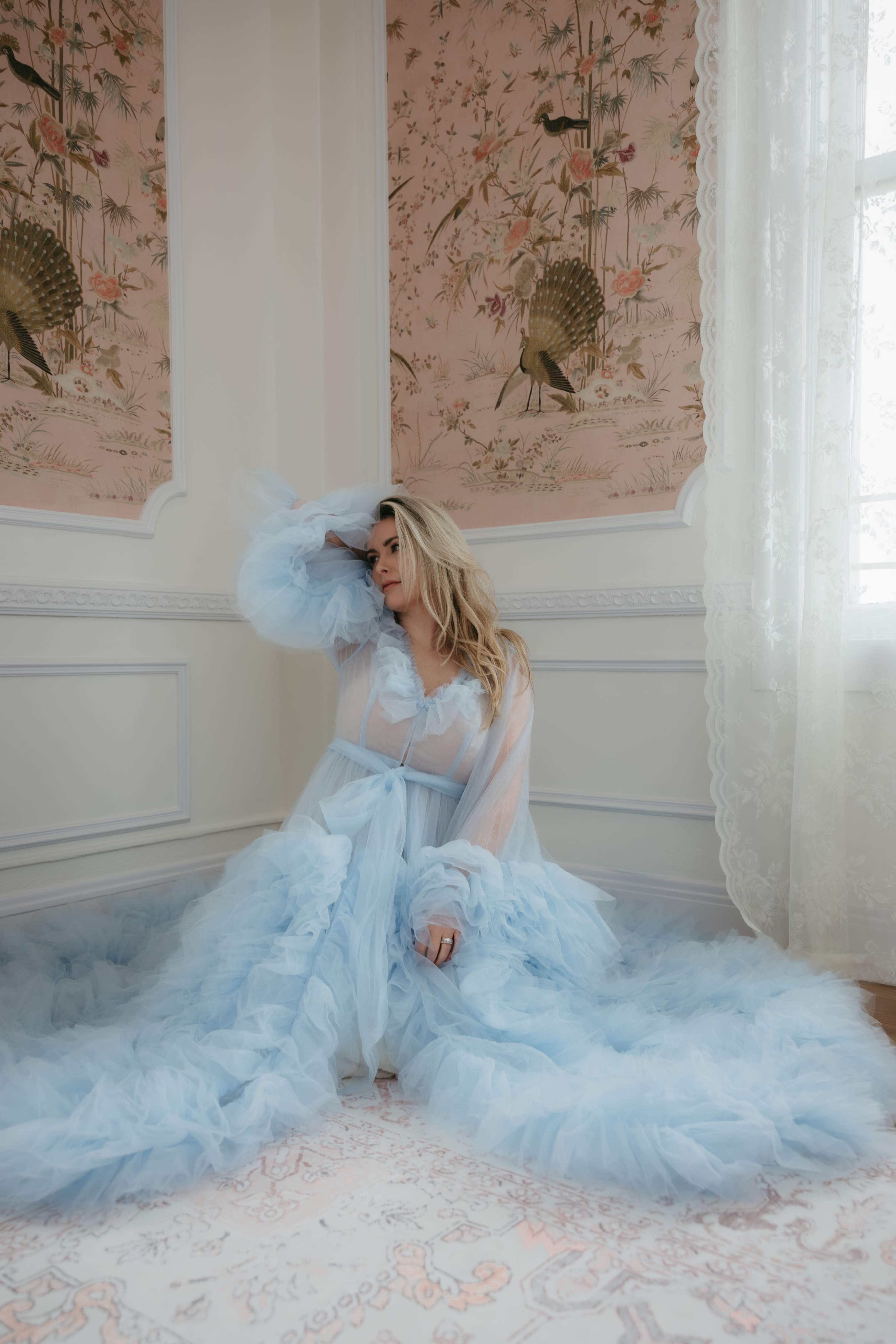 A woman in a light blue, ruffled gown sits against a floral wallpapered wall, with sunlight streaming through a nearby window.