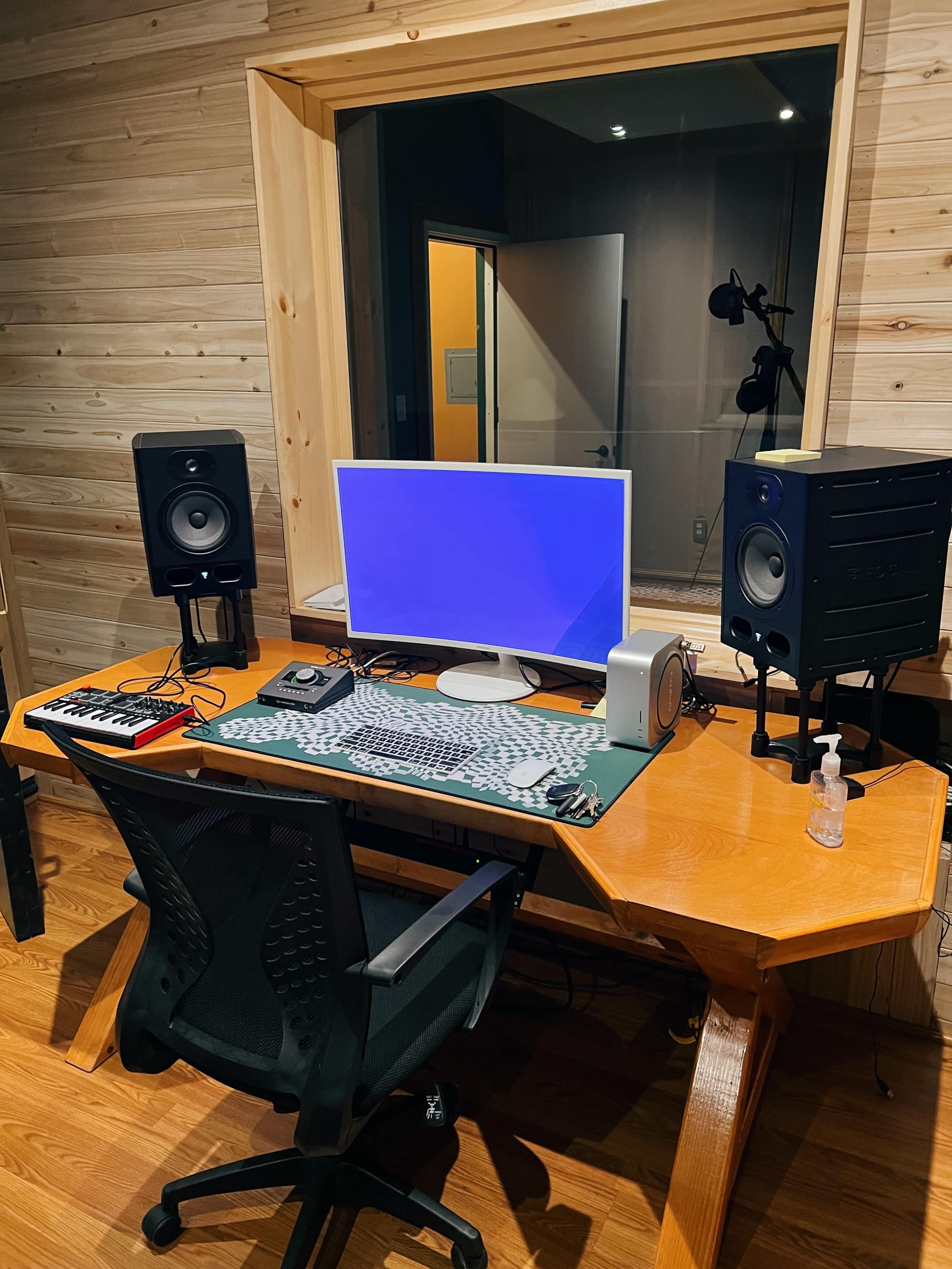 A wooden desk with a computer monitor, audio speakers, a MIDI keyboard, and a hand sanitizer dispenser is set up in a recording studio.