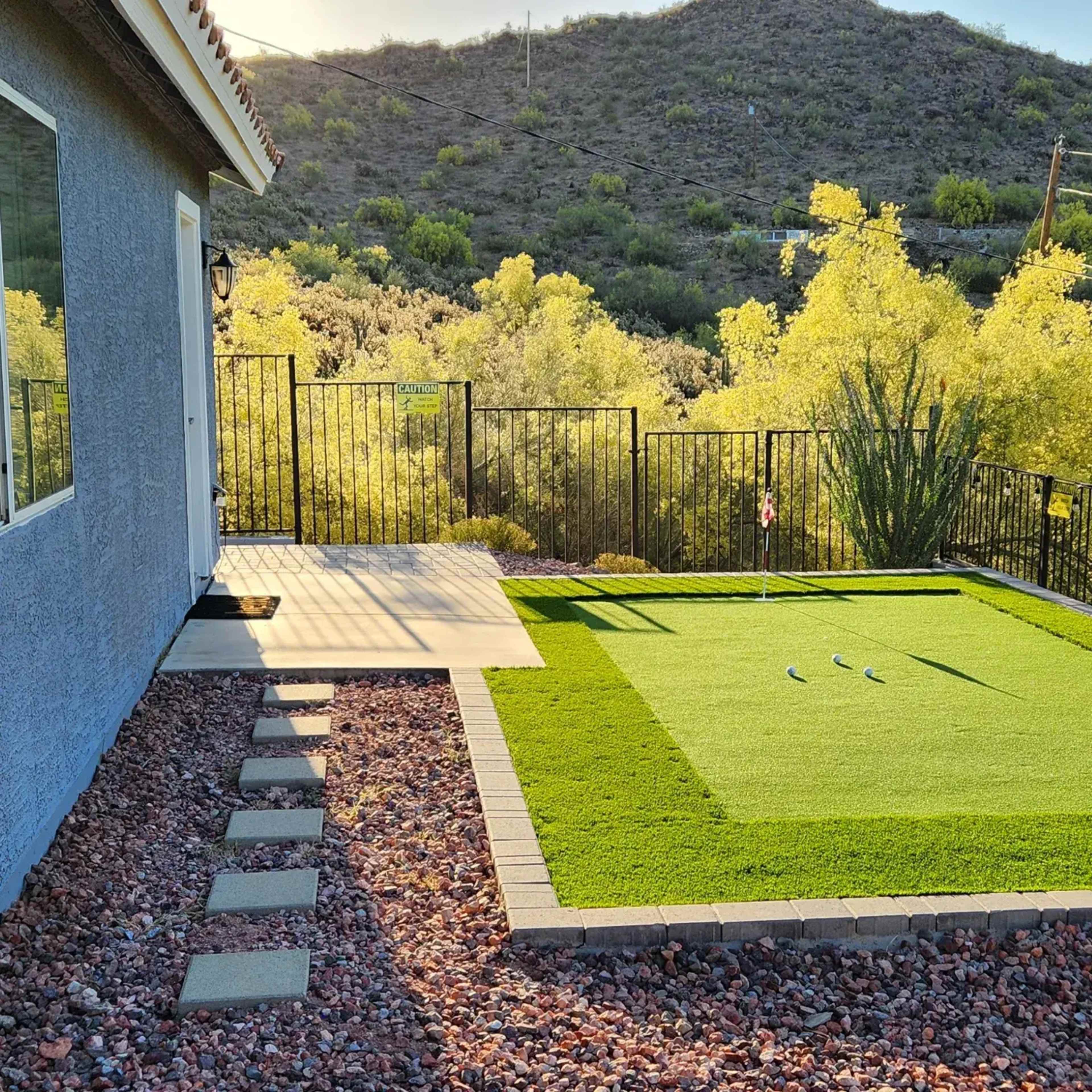 A well-maintained artificial putting green is situated next to a house, with a shaded patio area and a backdrop of green vegetation and hills.