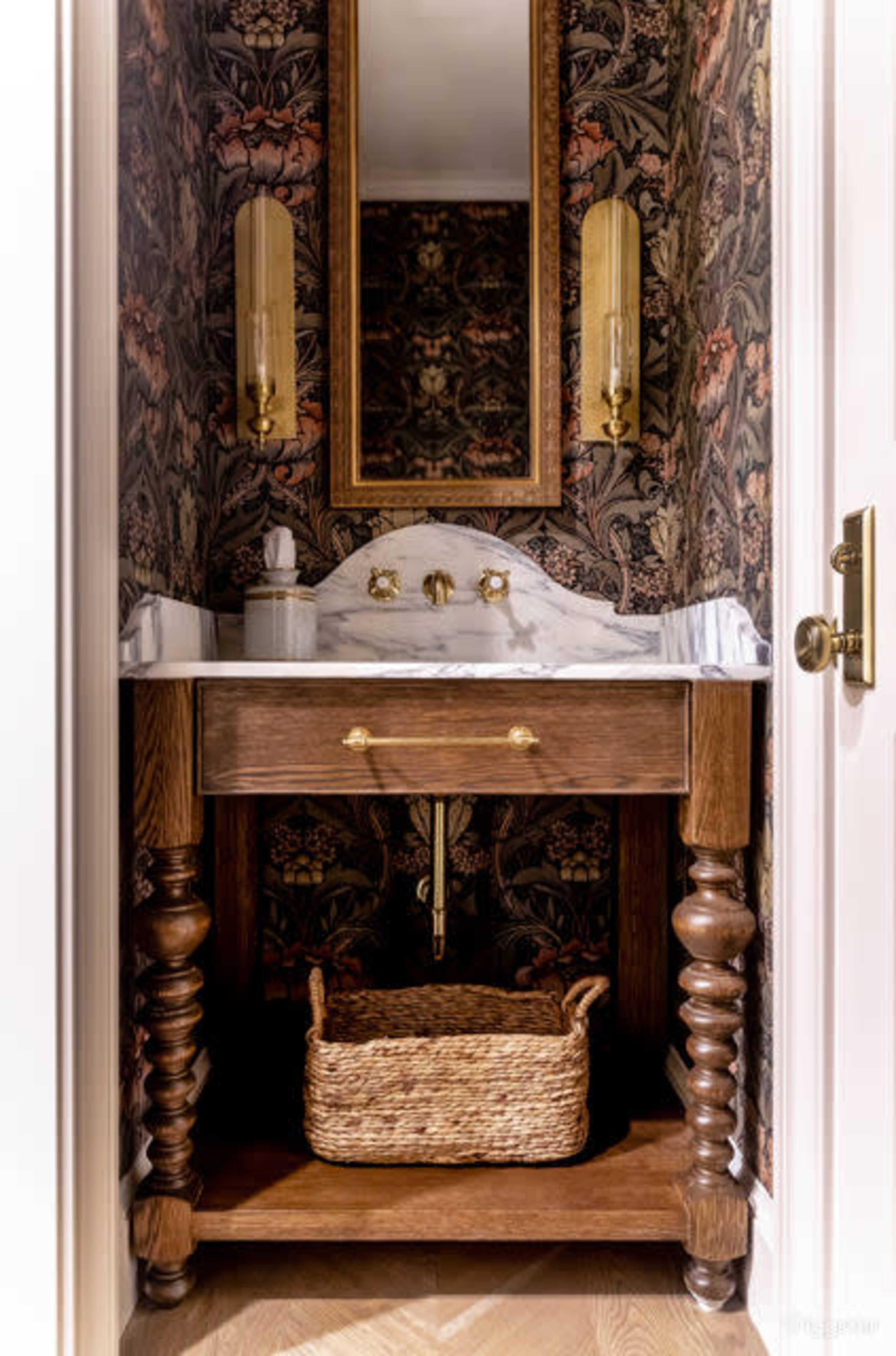 A small bathroom vanity with a marble countertop, two sink faucets, a mirror above, and decorative wallpaper featuring a floral pattern.