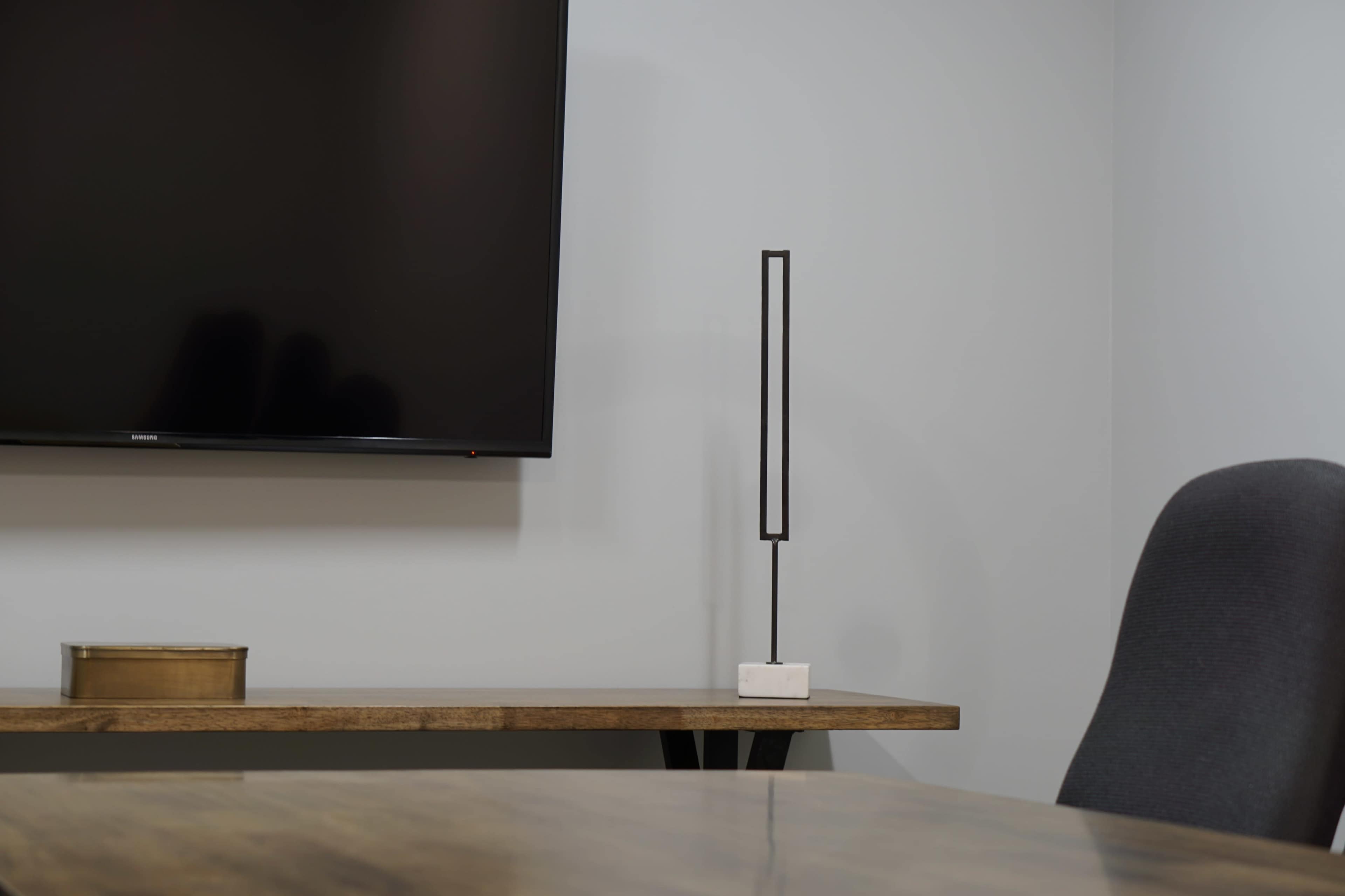 A modern workspace features a television mounted on a wall, a minimalist lamp on a wooden table, and a gray chair.