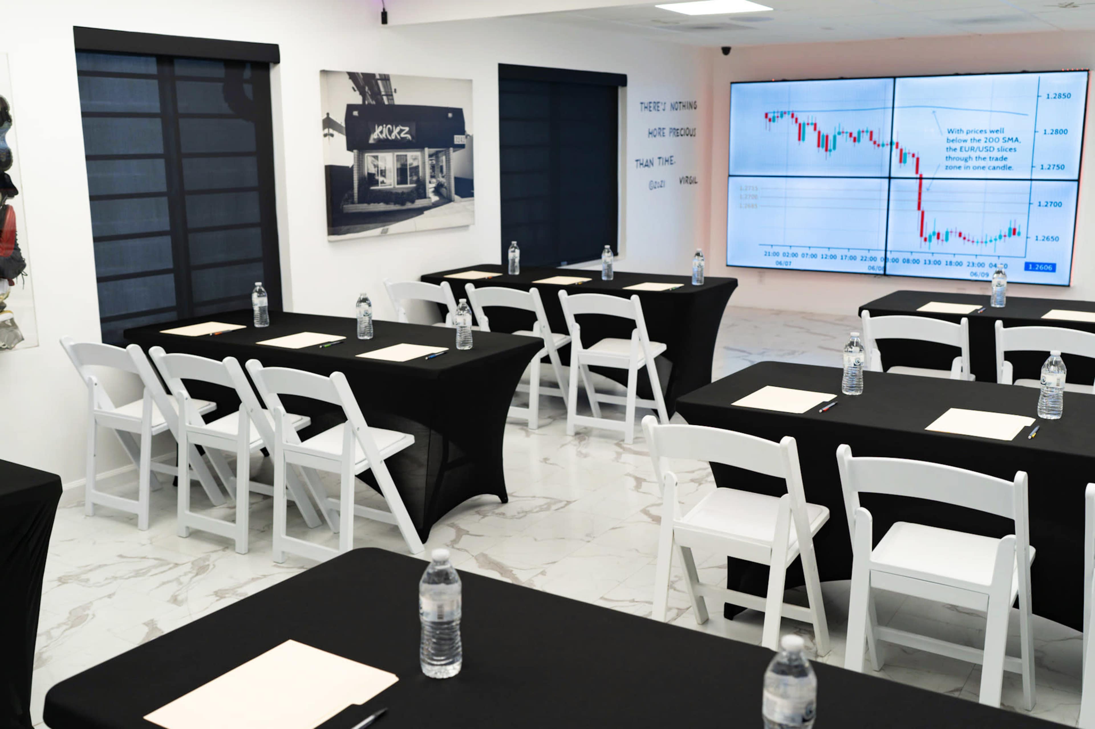 The image shows a modern conference room set up with several tables covered in black cloth, white chairs, and a large screen displaying a financial chart at the front.