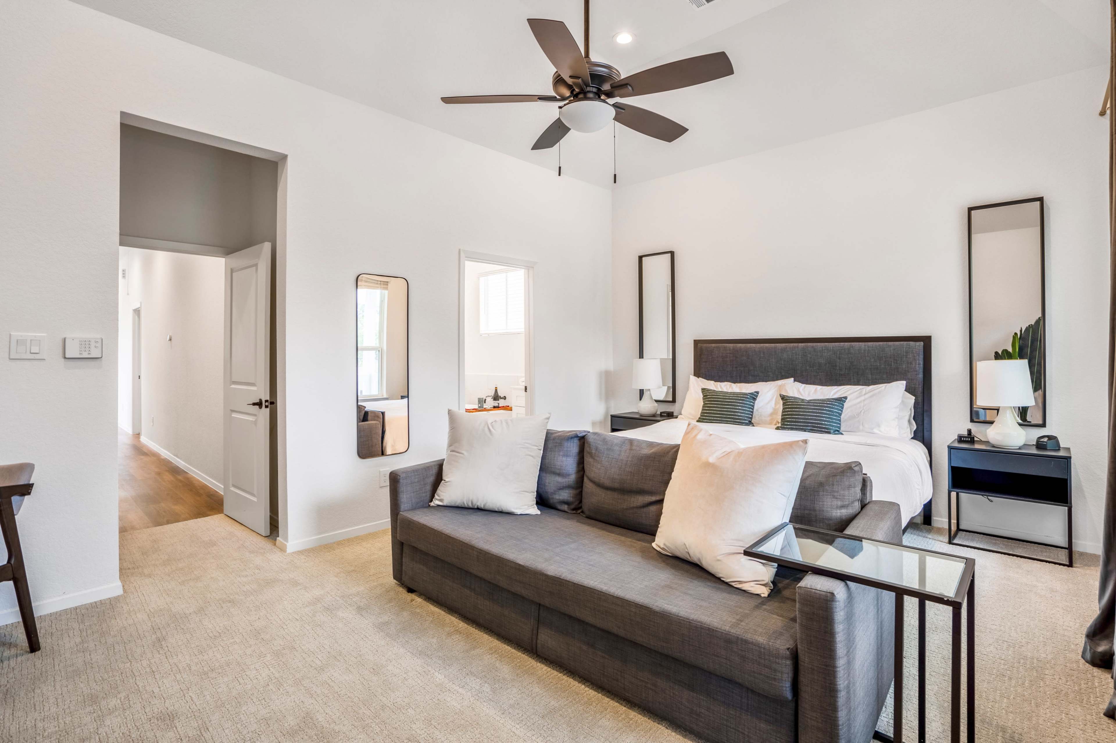 A neutral-toned bedroom features a bed with decorative pillows, a gray sofa with cushions, and a ceiling fan.