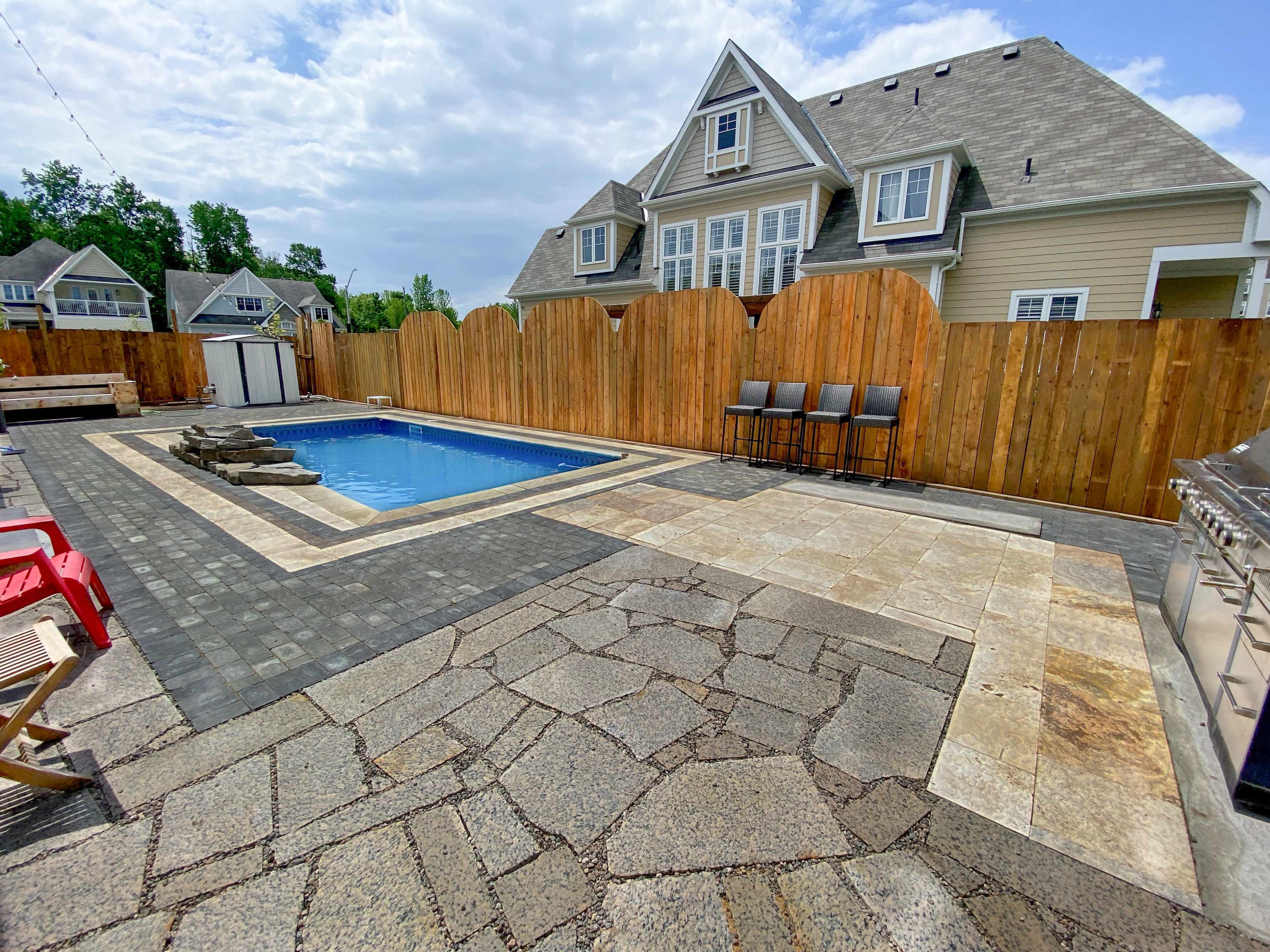 A rectangular swimming pool surrounded by a stone patio, with a wooden fence and a house in the background.