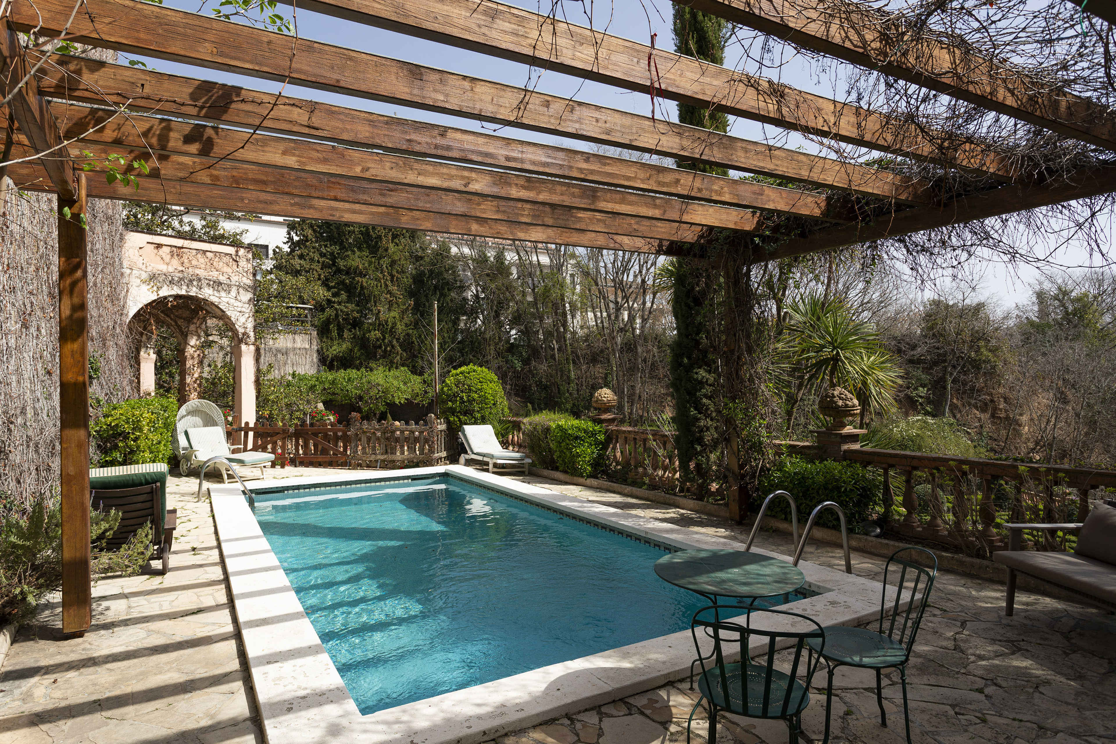 A rectangular swimming pool is surrounded by a stone patio, wooden pergola, and landscaped greenery.