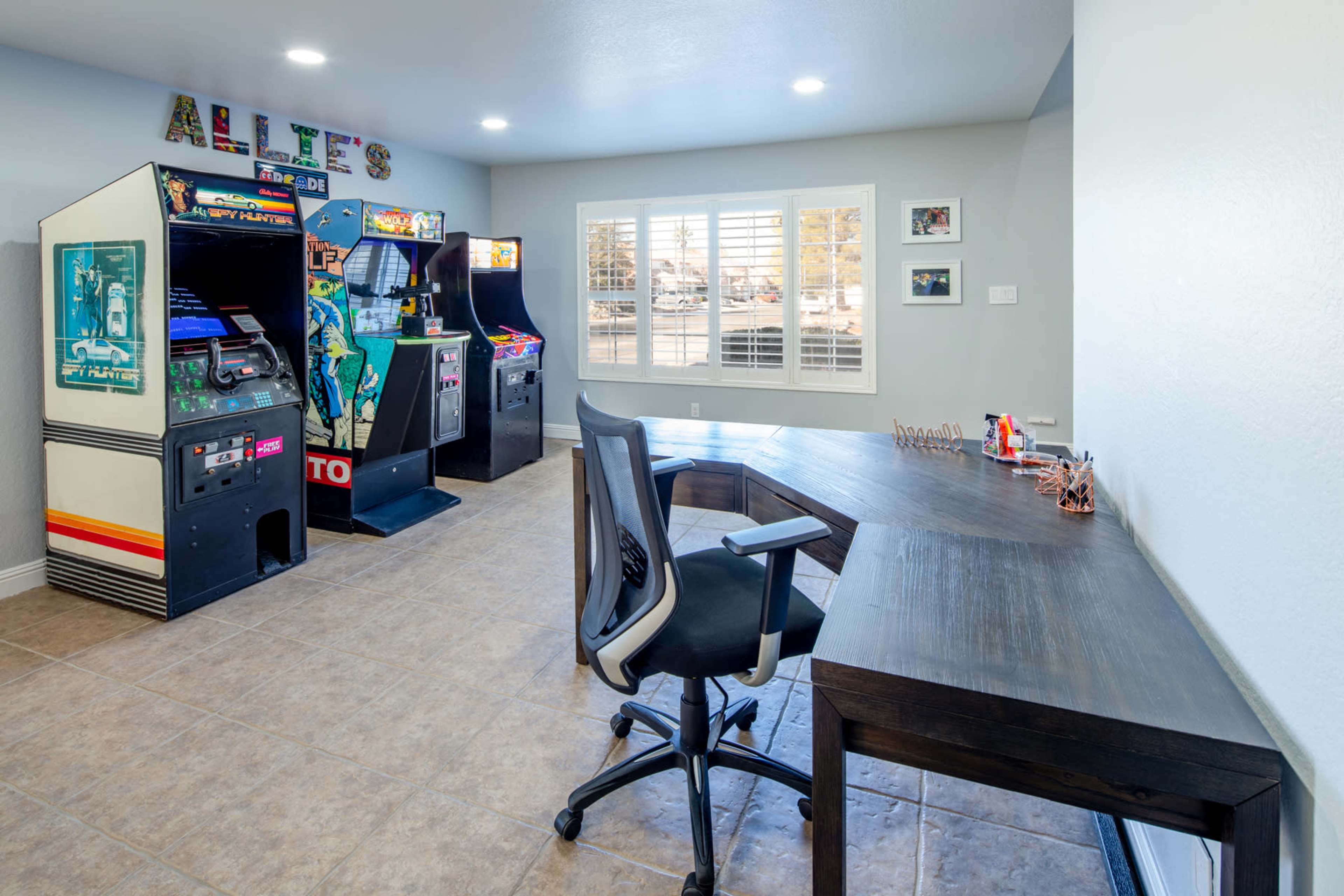 A modern home office featuring a large desk and two vintage arcade machines against the wall.