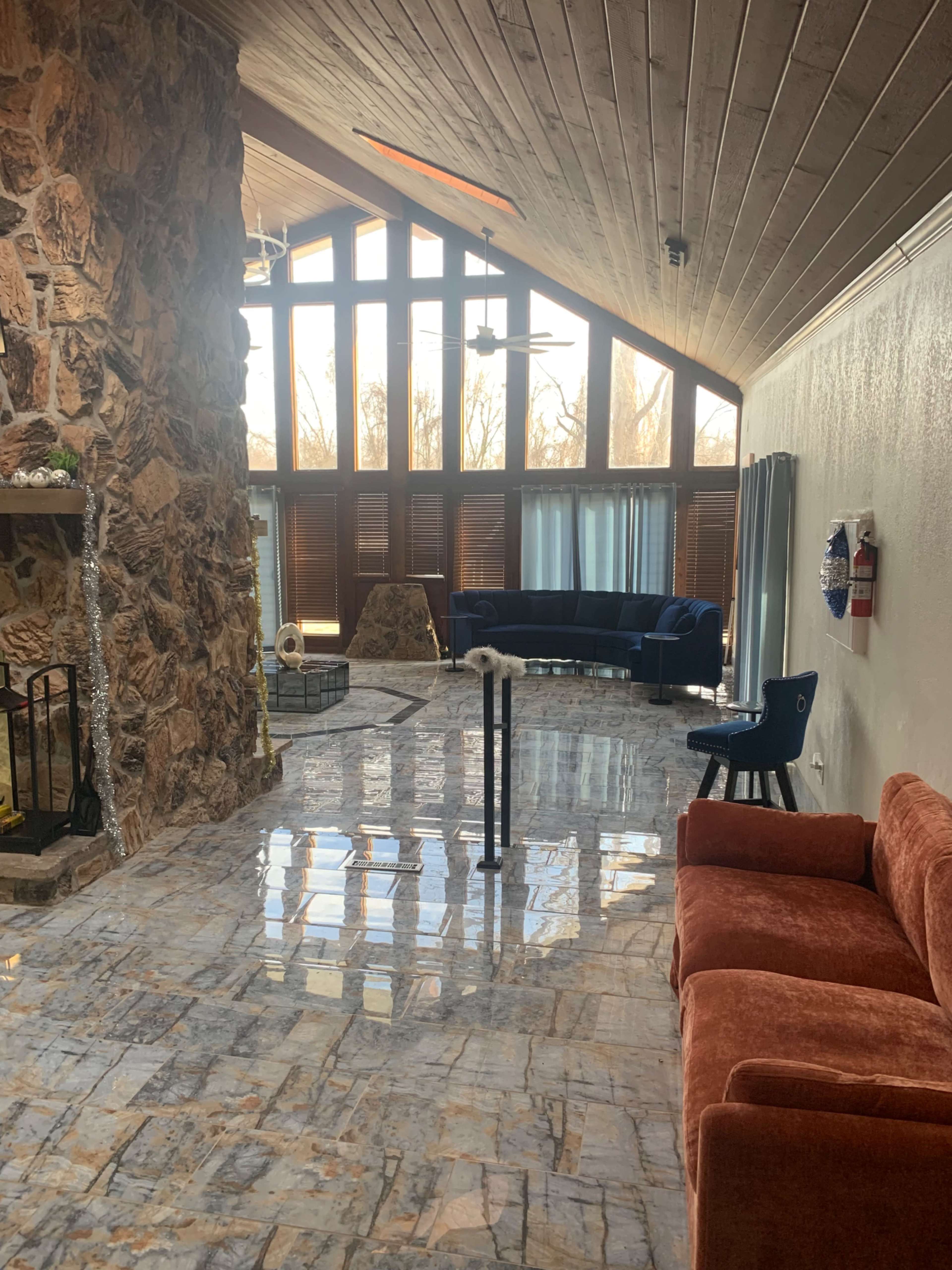 The image shows a spacious living room with a stone fireplace, large windows allowing natural light, and modern furniture including a blue sofa and an orange couch.