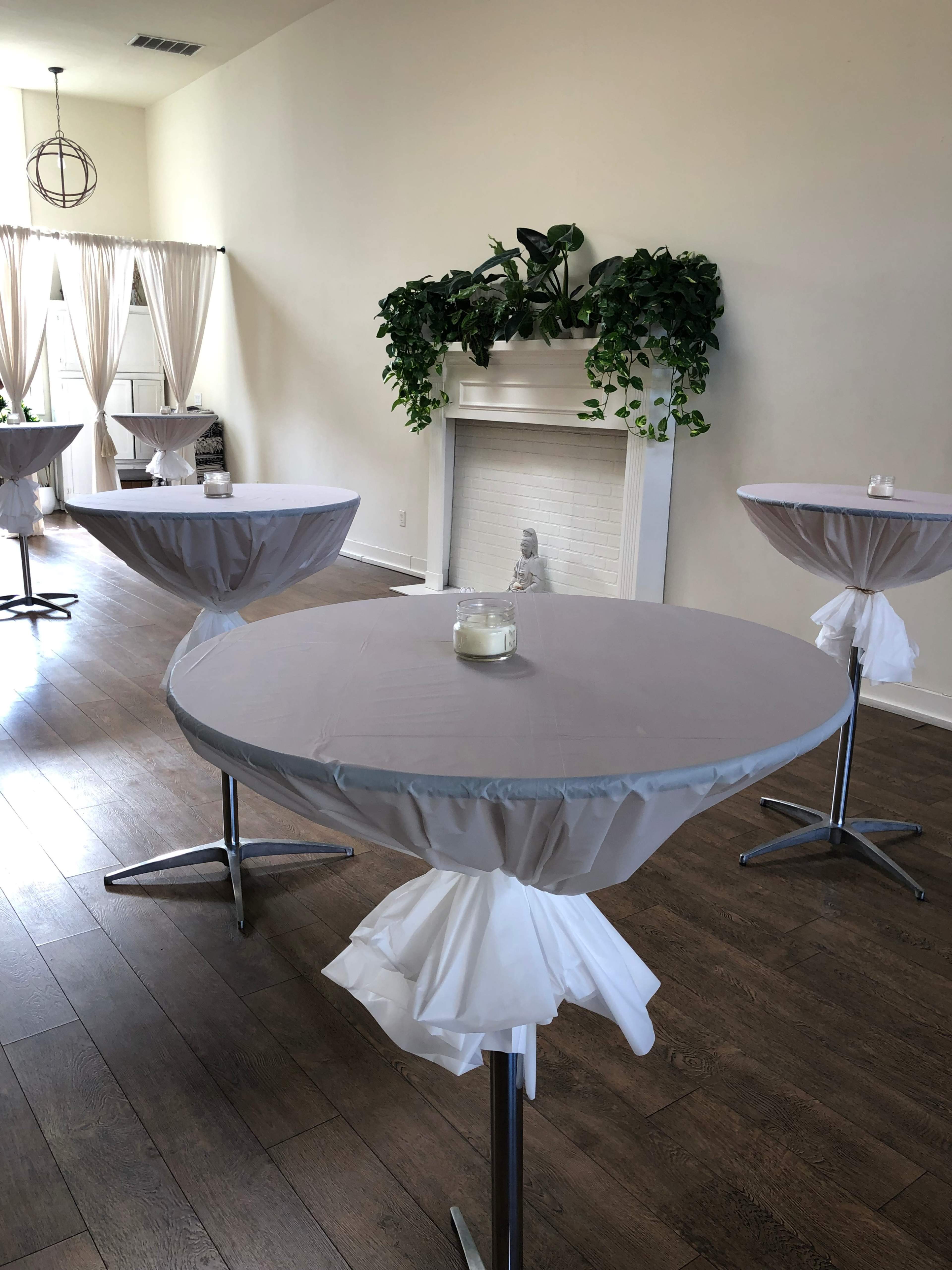 The image shows a well-lit event space with several tall, round tables covered in gray tablecloths and decorated with white accents, alongside a fireplace adorned with greenery.