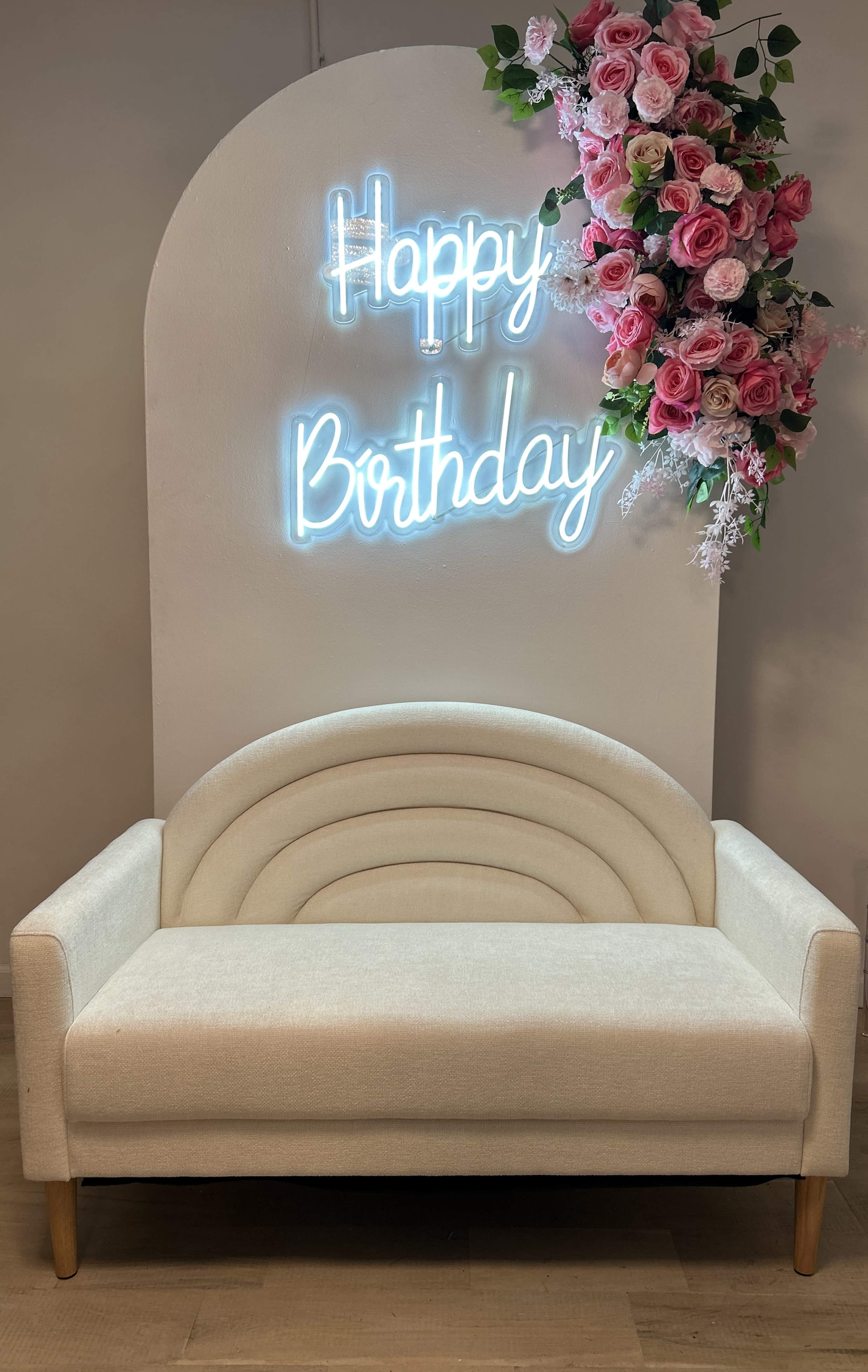 A lighted sign reading "Happy Birthday" is mounted on a wall adorned with pink roses, above a beige, curved-back sofa.