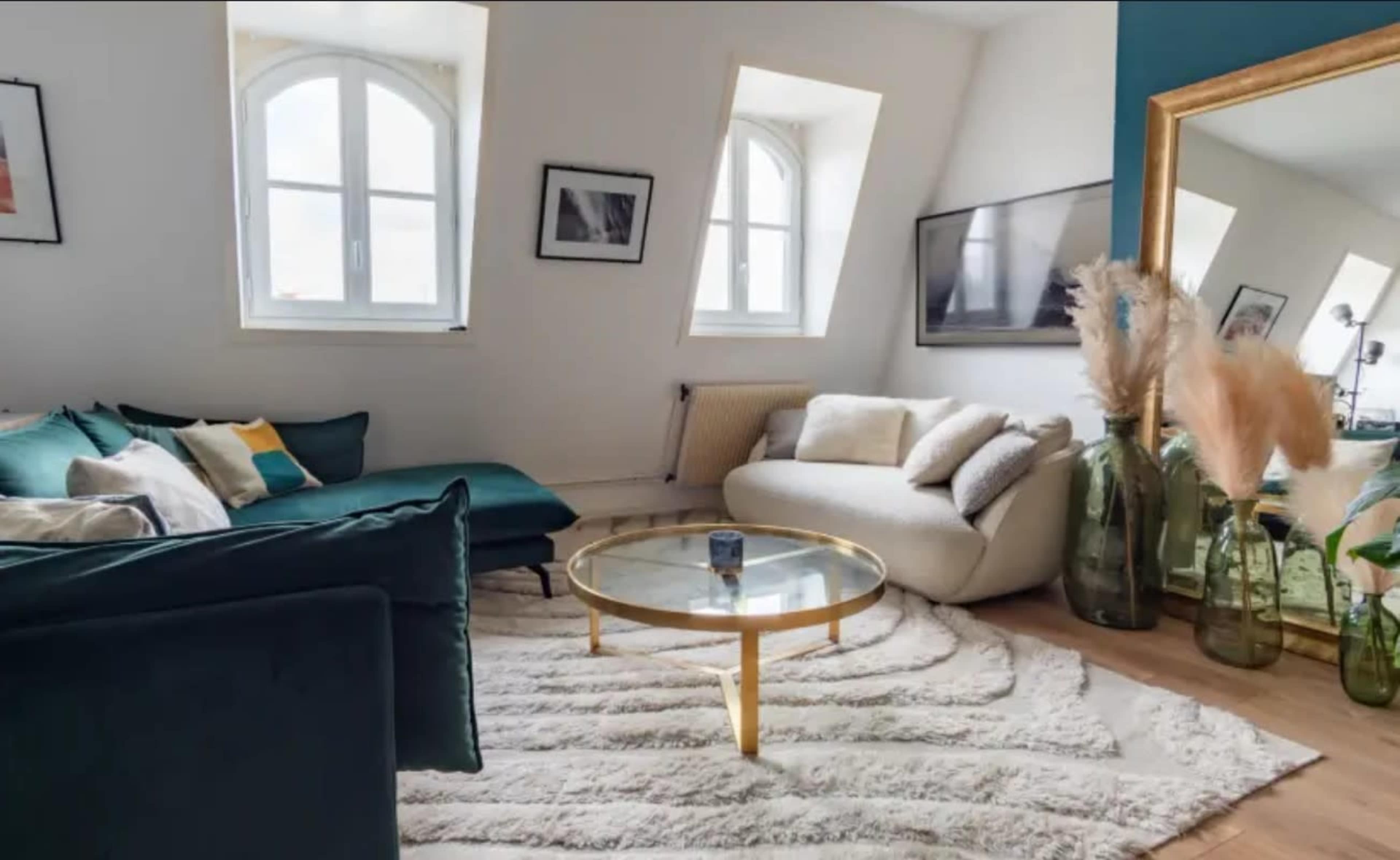 The image shows a modern living room with a green sectional sofa, a white curved couch, a circular glass coffee table, and decorative vases alongside a plush white rug.