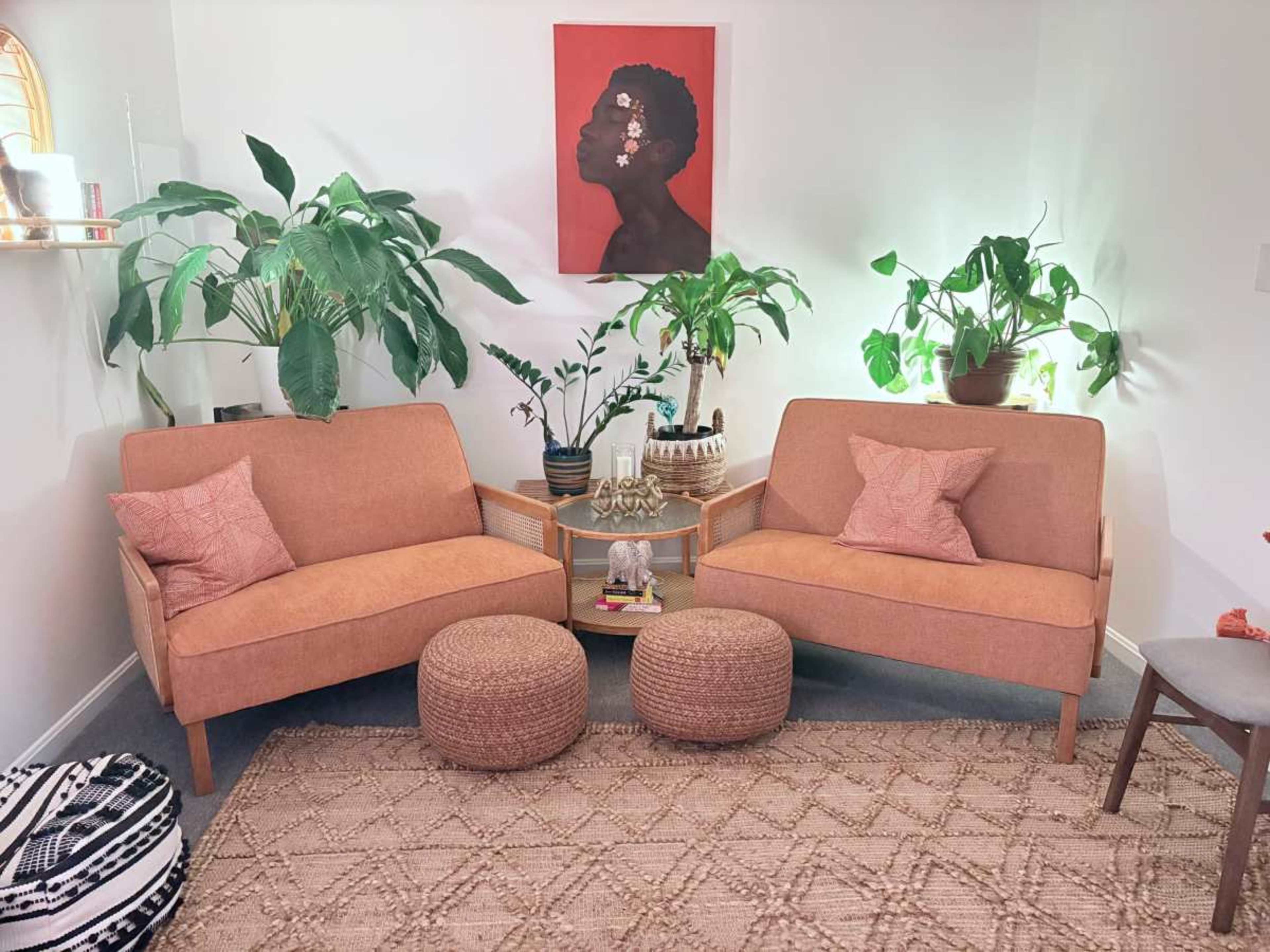 The image shows a cozy living room setup with two orange sofas, green plants, a round woven rug, and a large wall art piece depicting a profile portrait.