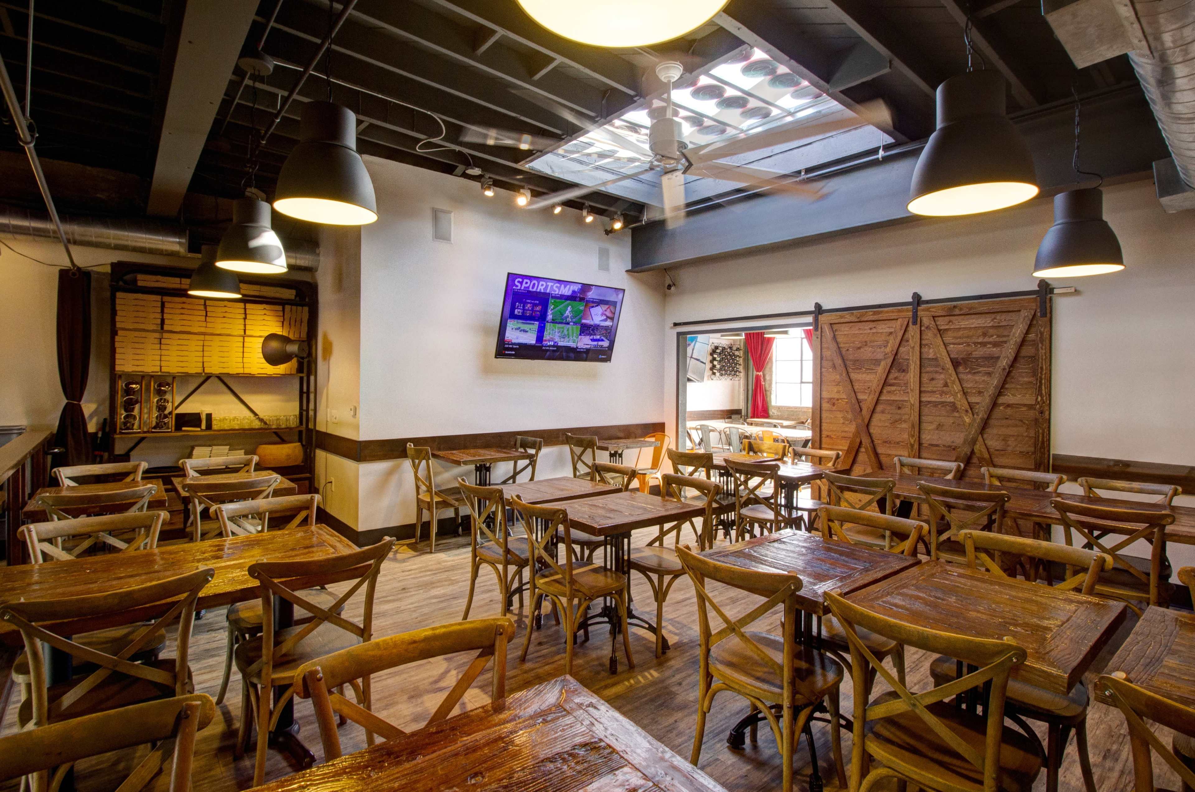 The image shows a spacious restaurant with wooden tables and chairs, featuring a TV on the wall and a rustic sliding door in the background.