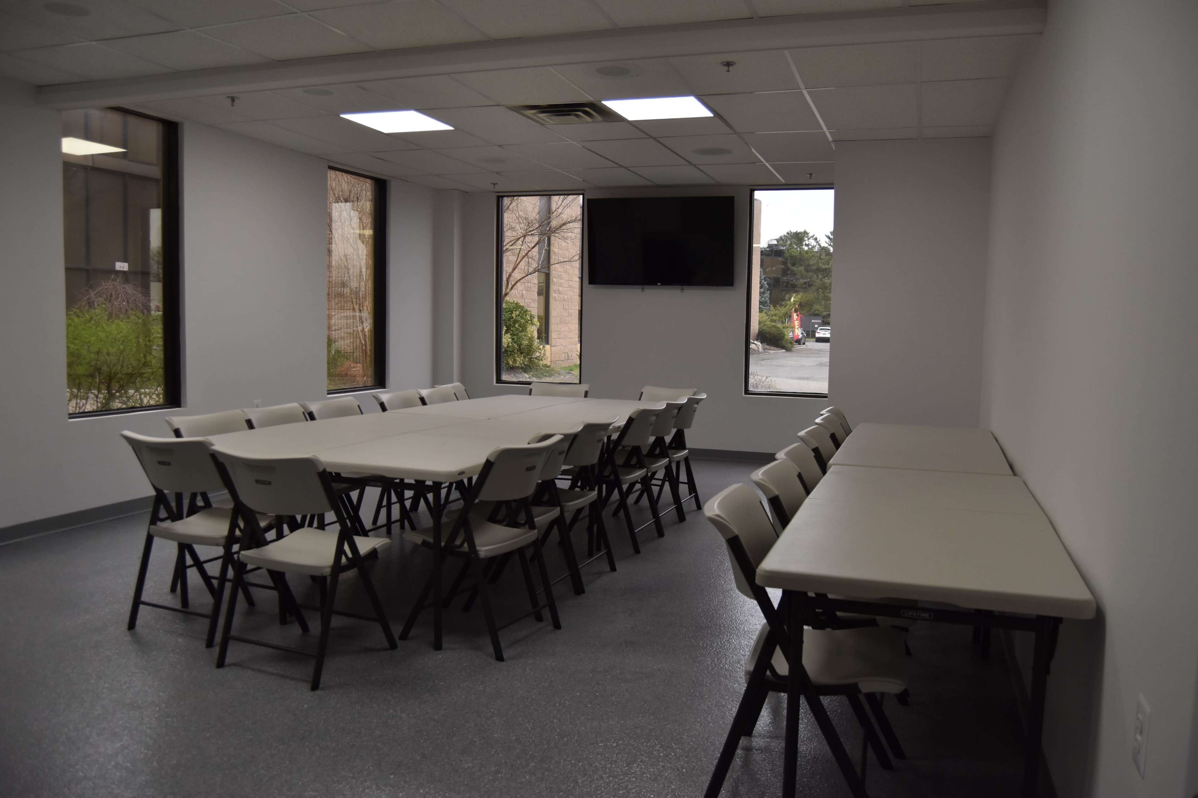 A meeting room features a large rectangular table surrounded by chairs, with large windows on two sides and a monitor mounted on the wall.