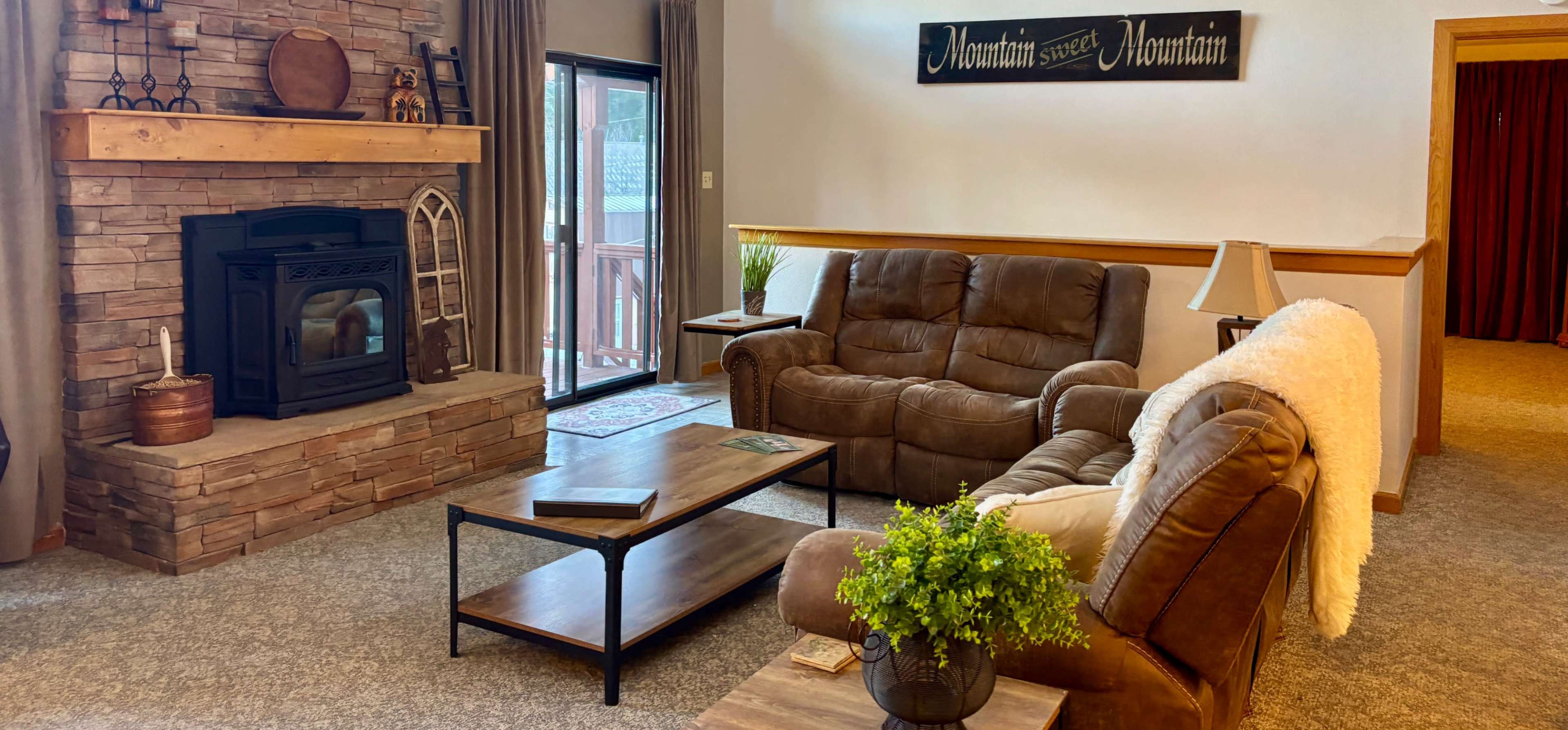 The image shows a living room with a stone fireplace, two sofas, a coffee table, and a large window overlooking a deck.