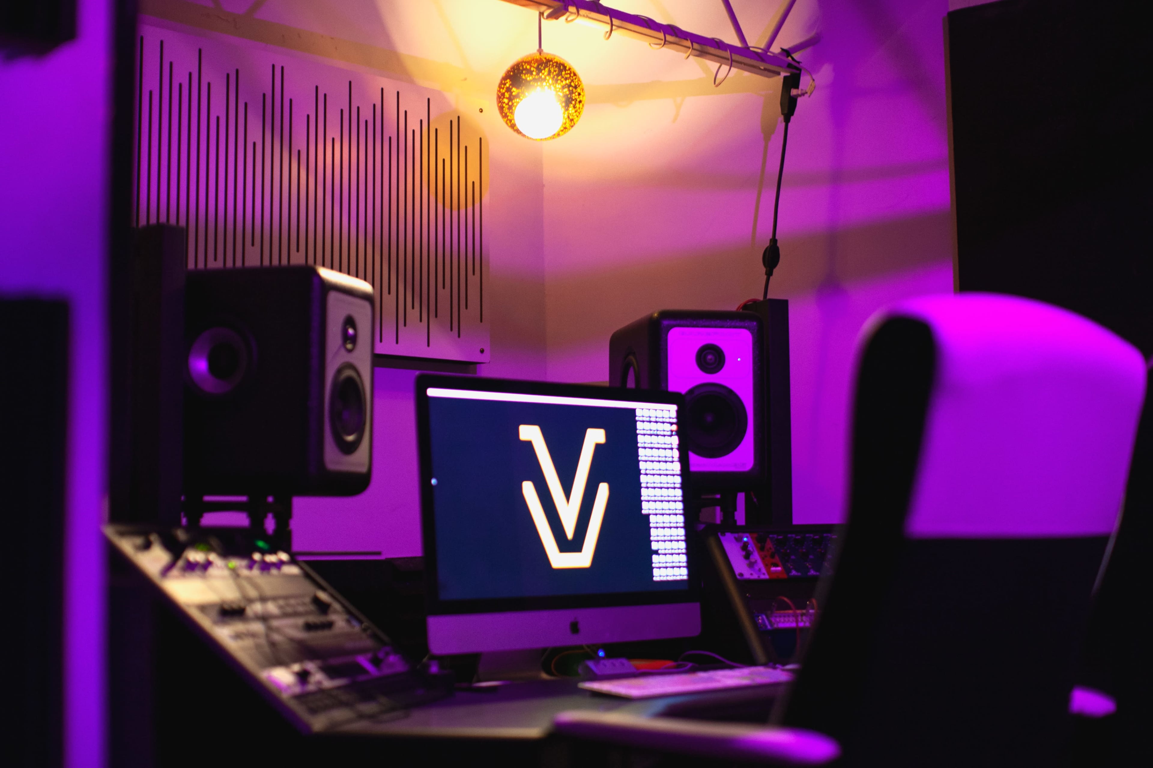 A music studio scene featuring a computer monitor displaying a logo, audio speakers, and a dim overhead light.