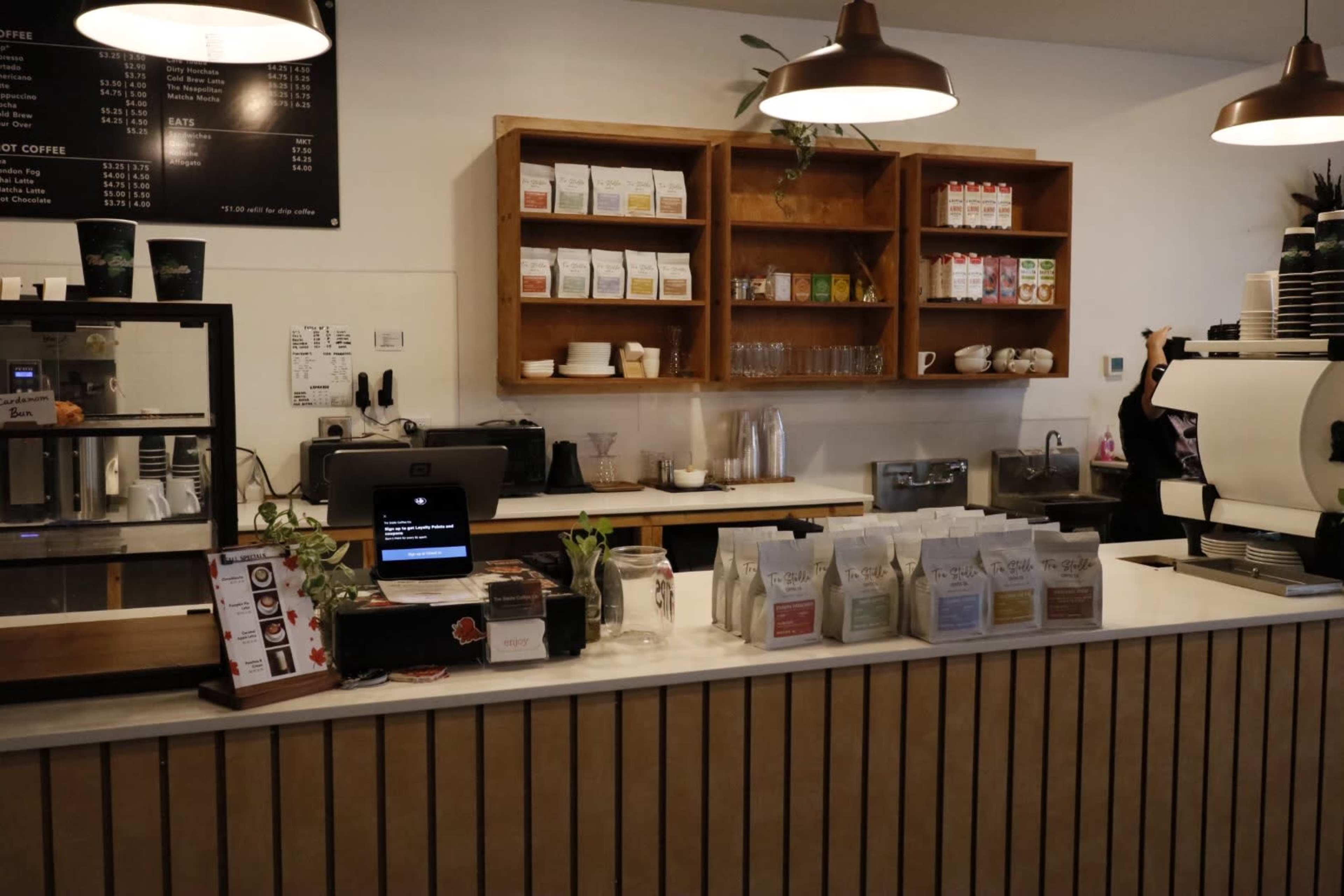 The image shows a coffee shop counter with a coffee machine, display of coffee bags, and shelves filled with various drink options.