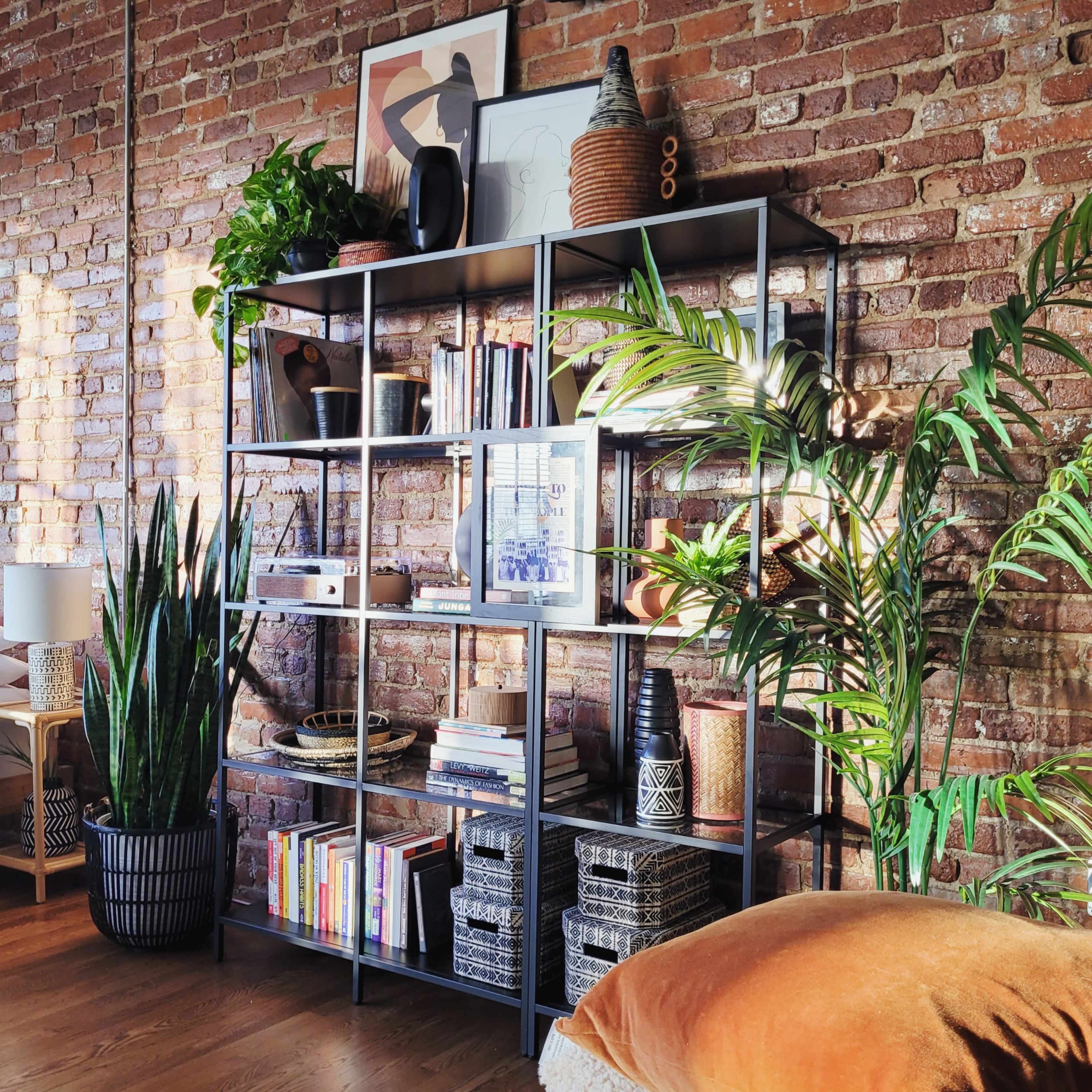 A modern bookshelf filled with books, decor, and plants stands against a brick wall in a well-lit room.