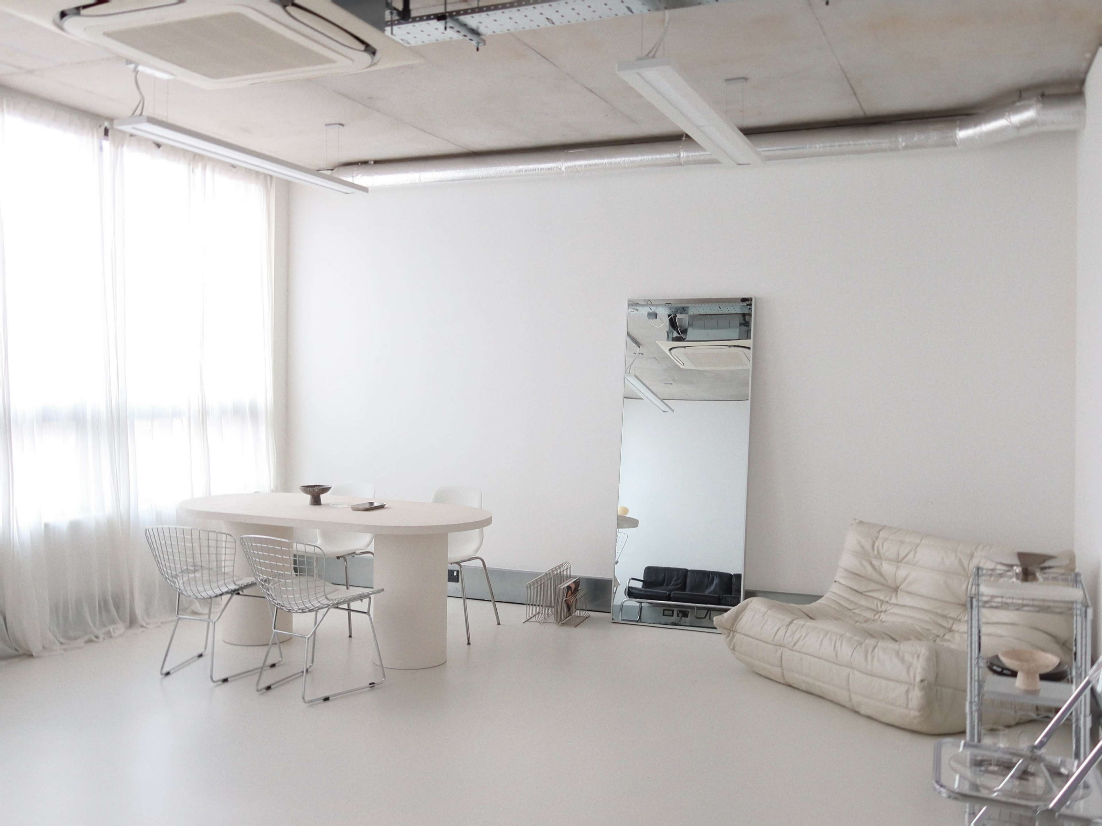 The image features a minimalist interior with a round table surrounded by wire chairs, a large mirror, and a cream-colored lounge chair in a spacious room with white walls and natural light.