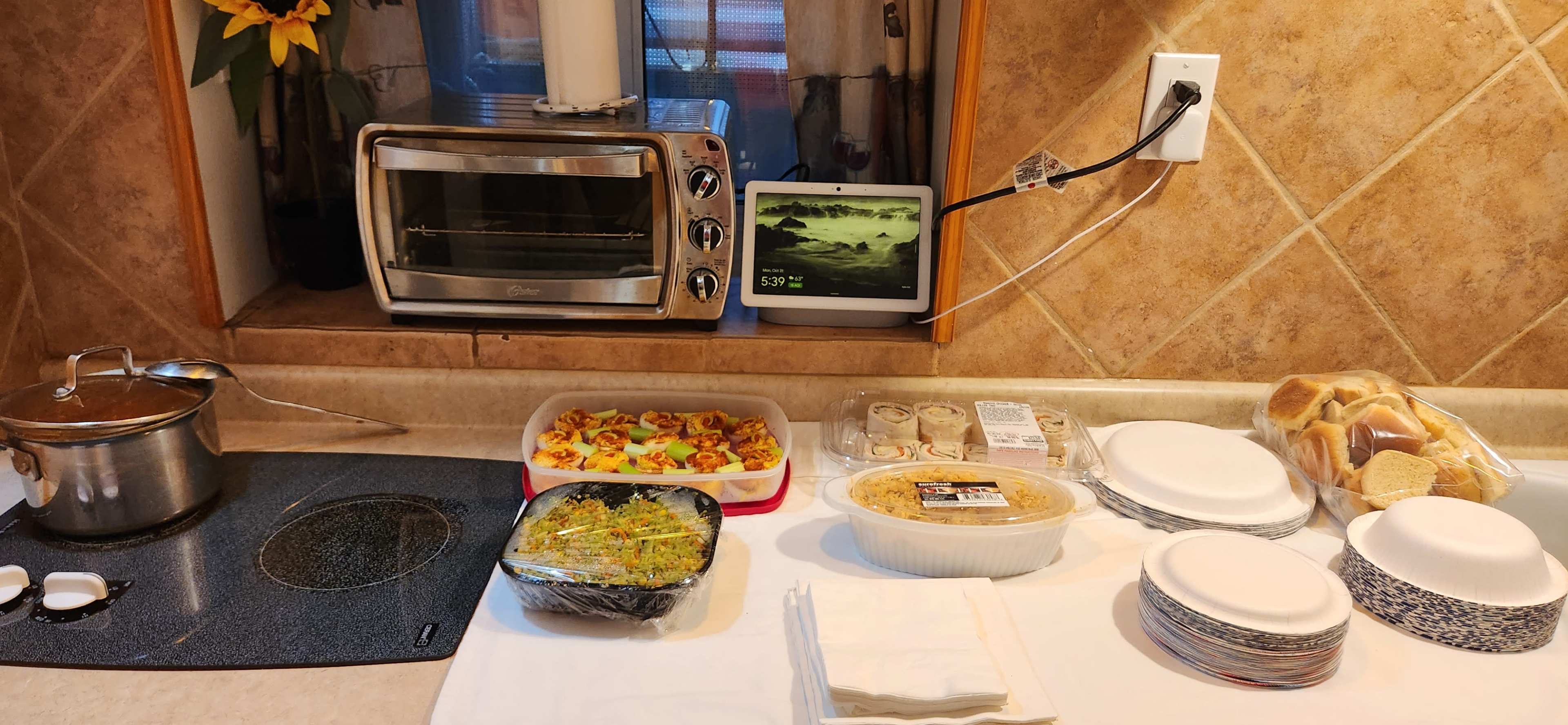 A kitchen counter displays a variety of food items, including salad, main dishes, and baked goods, alongside a toaster oven and a digital display.