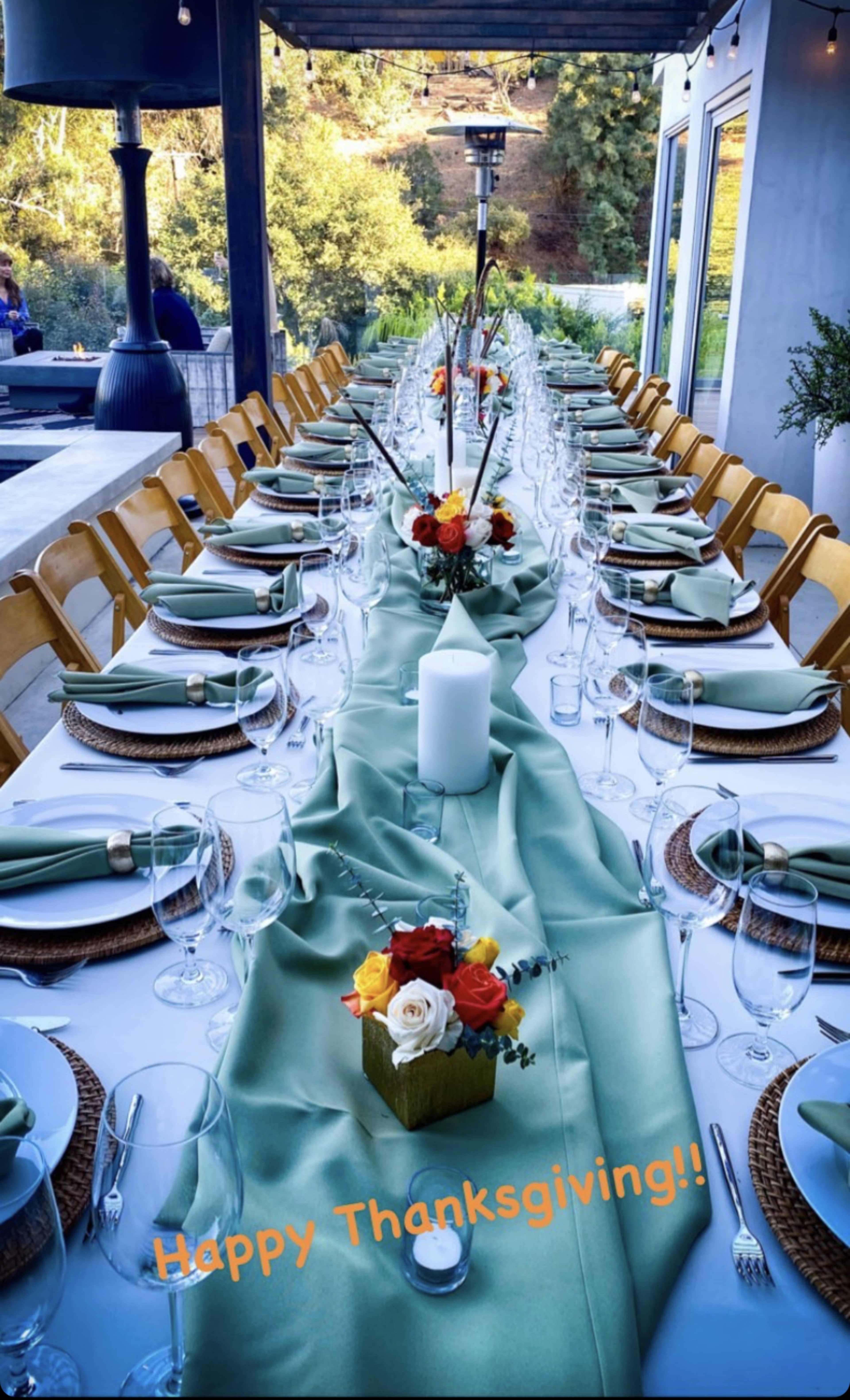 A long dining table is elegantly set with green linens, glassware, and floral centerpieces, ready for a Thanksgiving meal.
