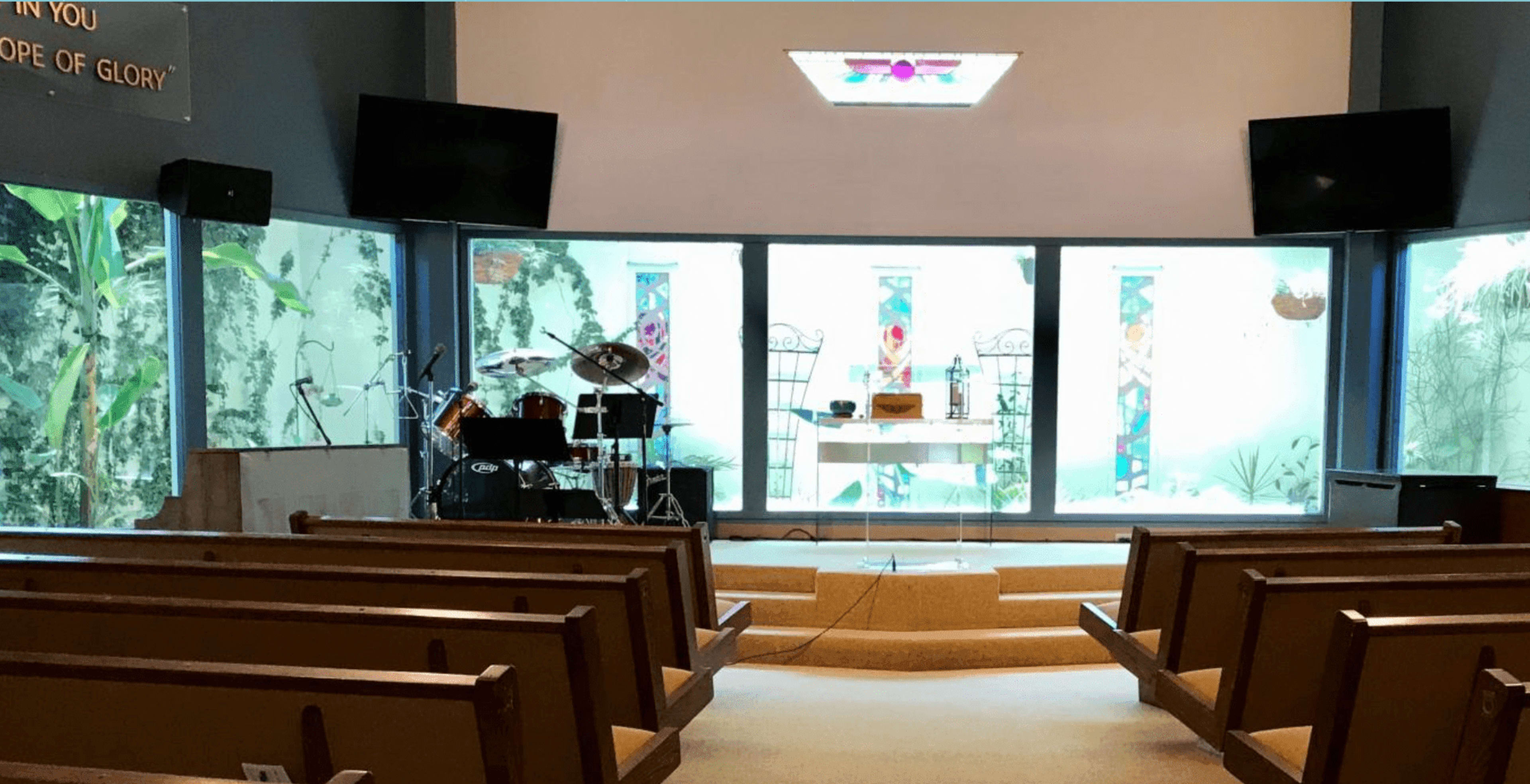 The interior of a church features rows of wooden pews facing a central stage area with musical instruments and large windows displaying greenery.