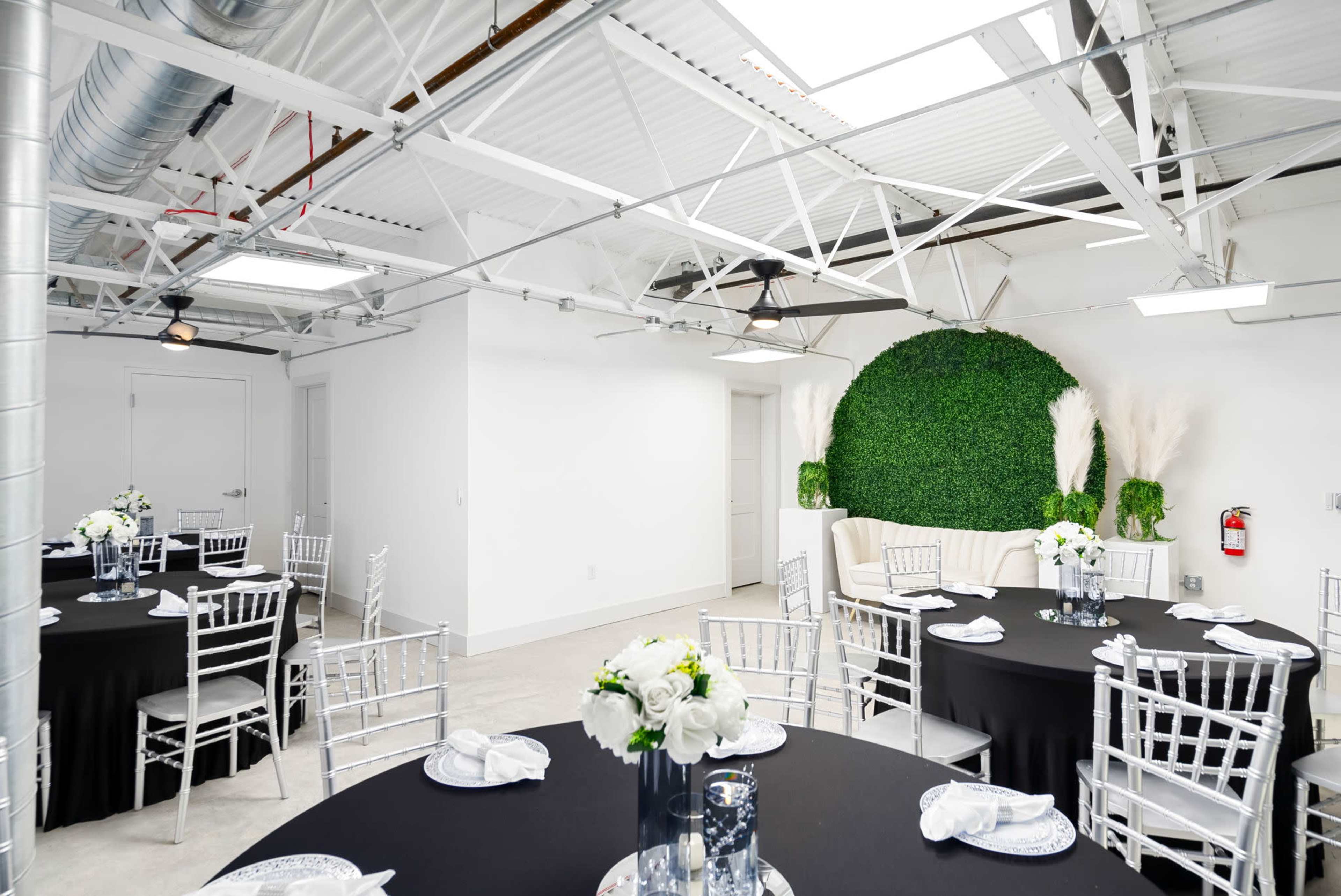 A modern event space featuring round tables with black tablecloths and silver chiavari chairs, accented by greenery on a wall and decorative floral arrangements.