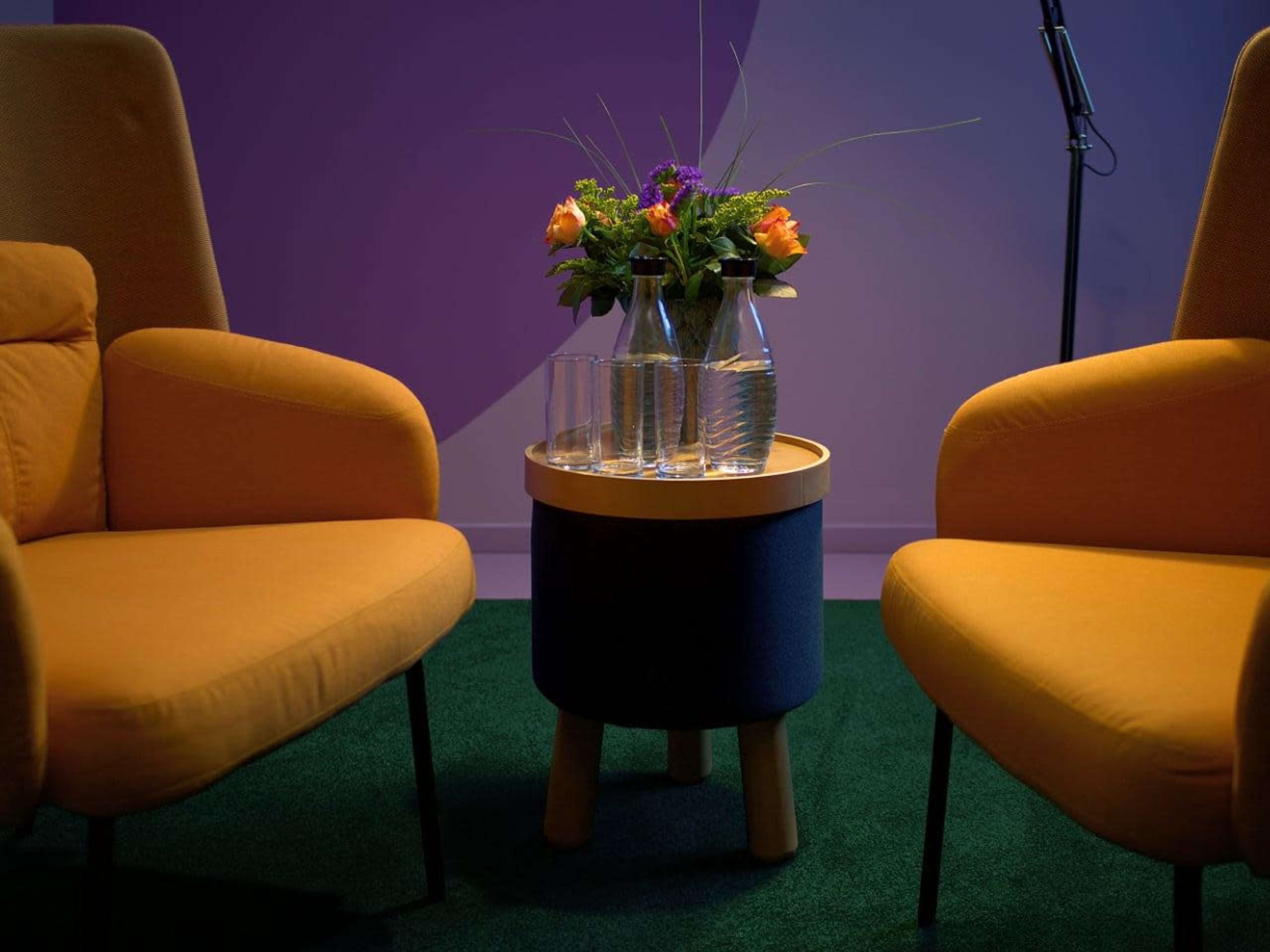 A small round table with a glass pitcher and two glasses sits between two yellow armchairs, accompanied by a vase of flowers.