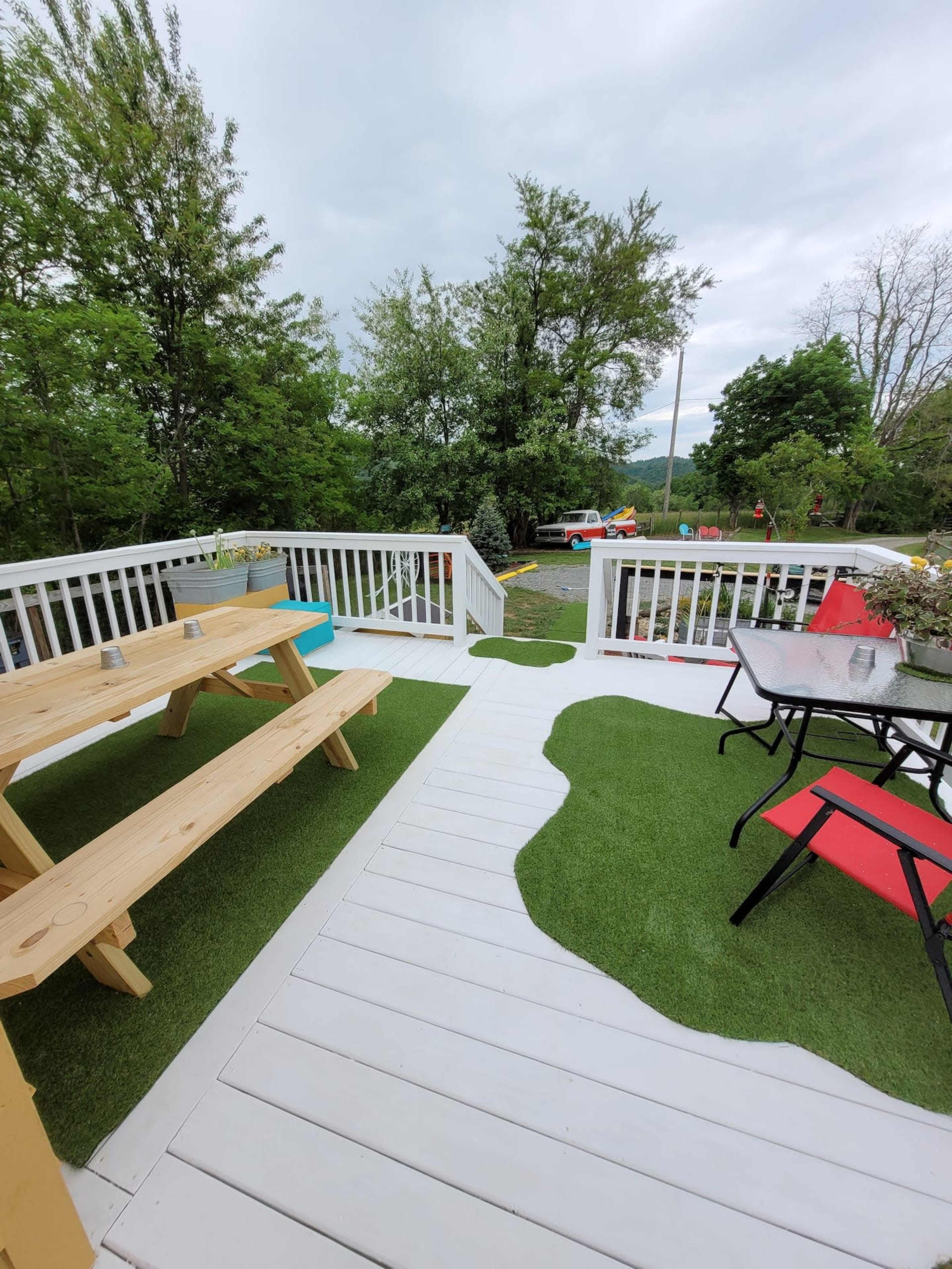 A deck with wooden picnic tables and black and red outdoor chairs, surrounded by green grass and trees.