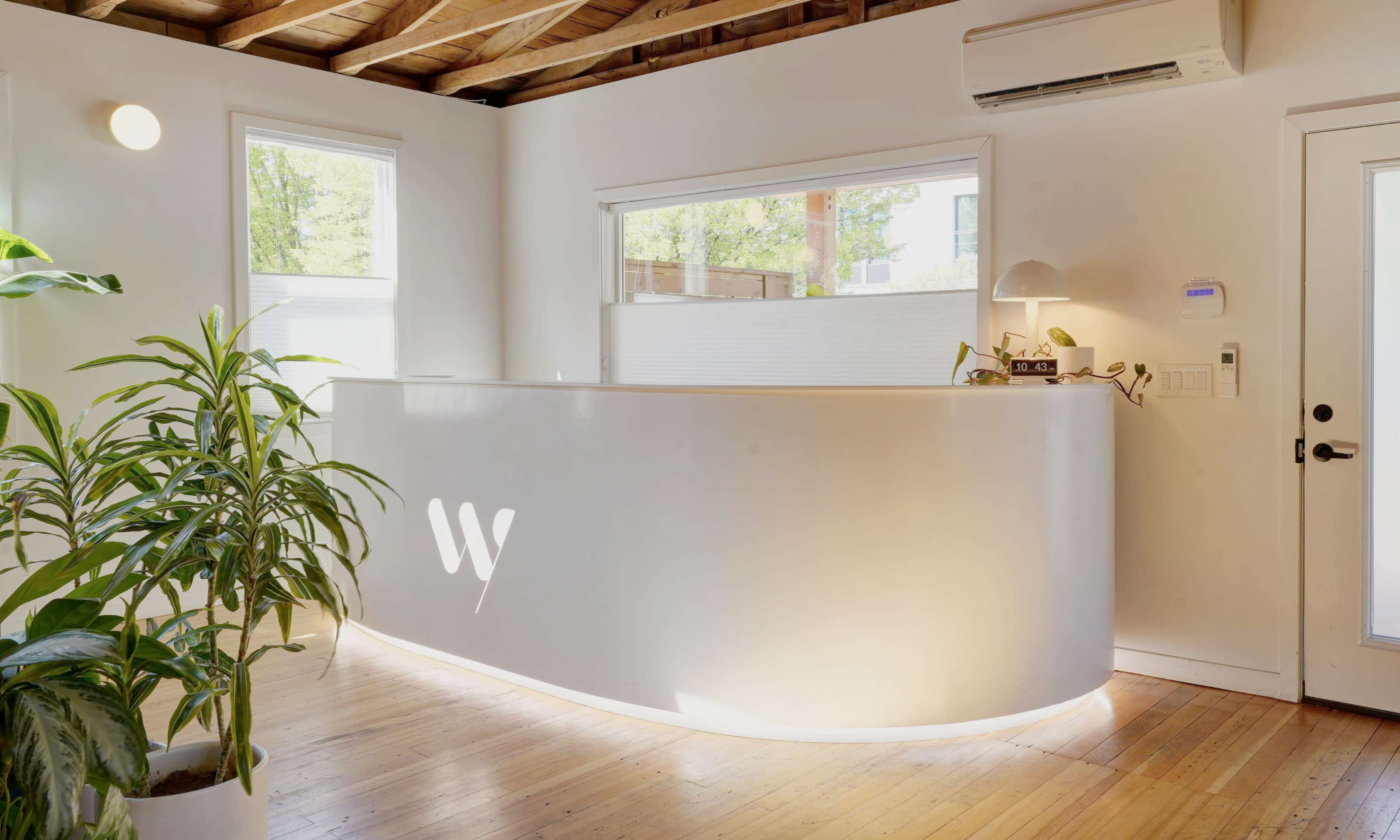 The image shows a modern reception area with a curved white desk, large windows allowing natural light, and a potted plant in a wooden-floored space.
