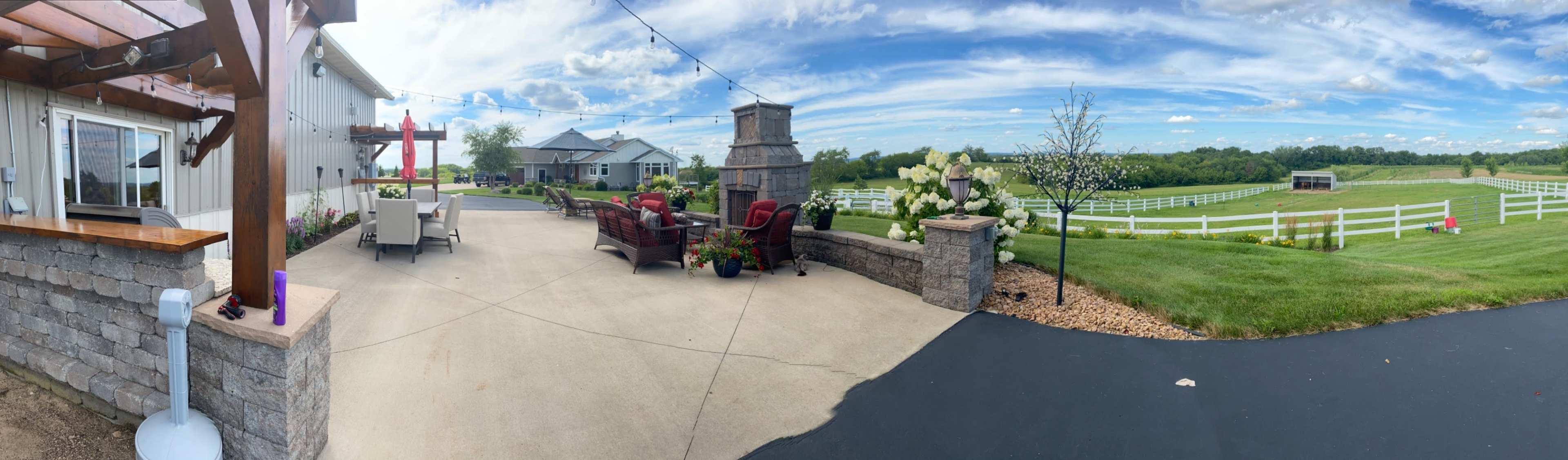 A panoramic view of a stone patio area with seating, surrounded by green fields and a white fence under a blue sky with clouds.