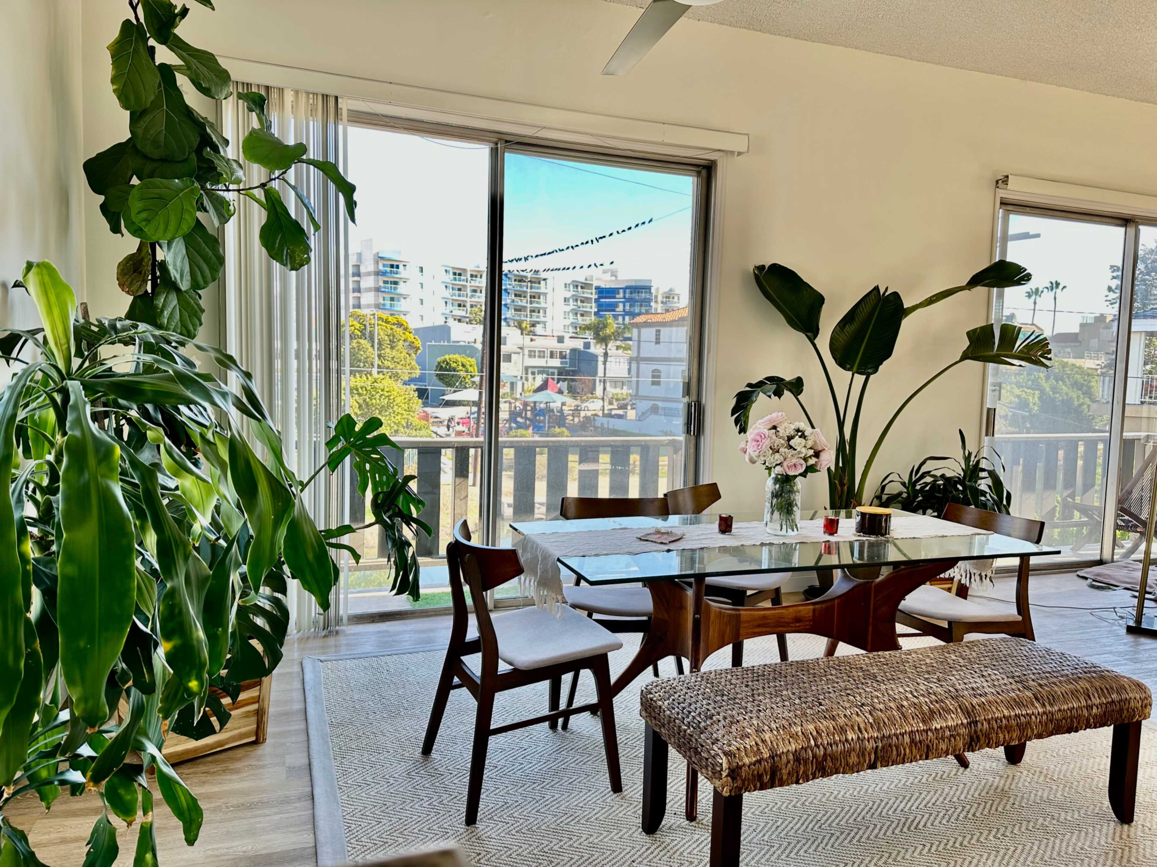 A bright dining area features a wooden table with chairs, surrounded by large plants and a view of the street through large windows.