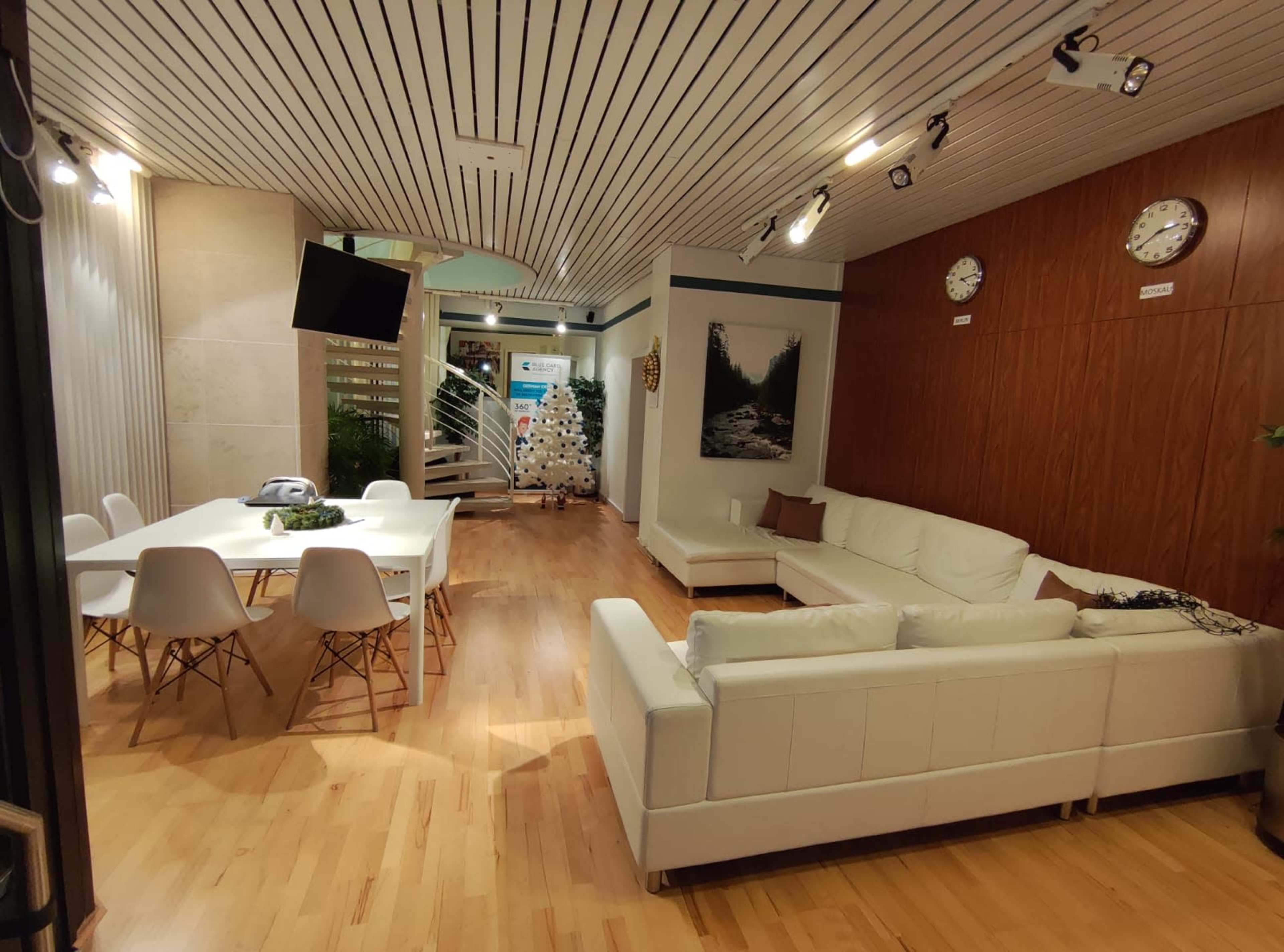 The image shows a modern office interior with a white sofa, a dining table, and a TV on the wall, featuring wooden and light-colored floors.