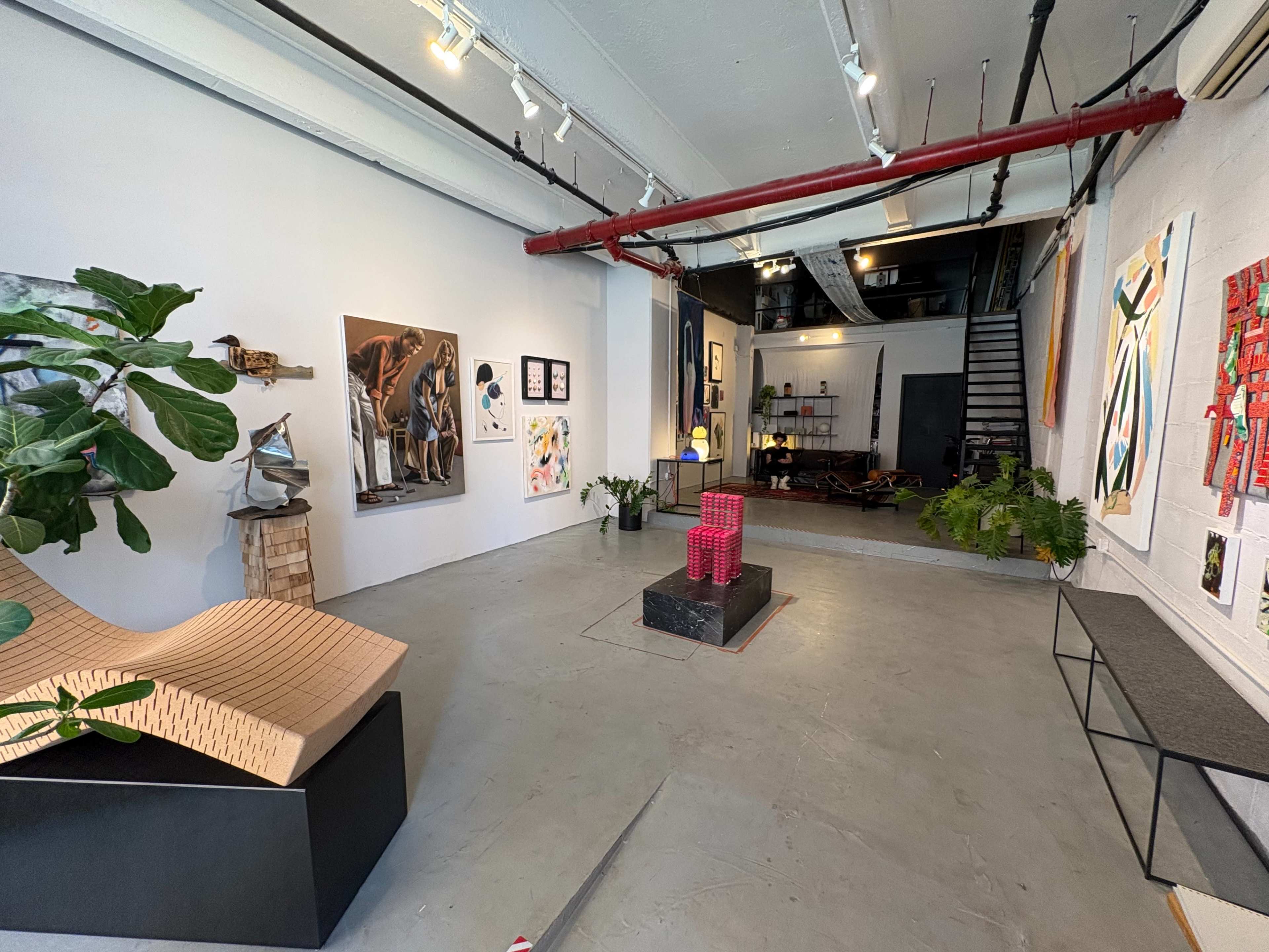 The image shows a spacious art gallery featuring various artworks on the walls, a unique wooden sculpture in the foreground, and a staircase leading to an upper area.