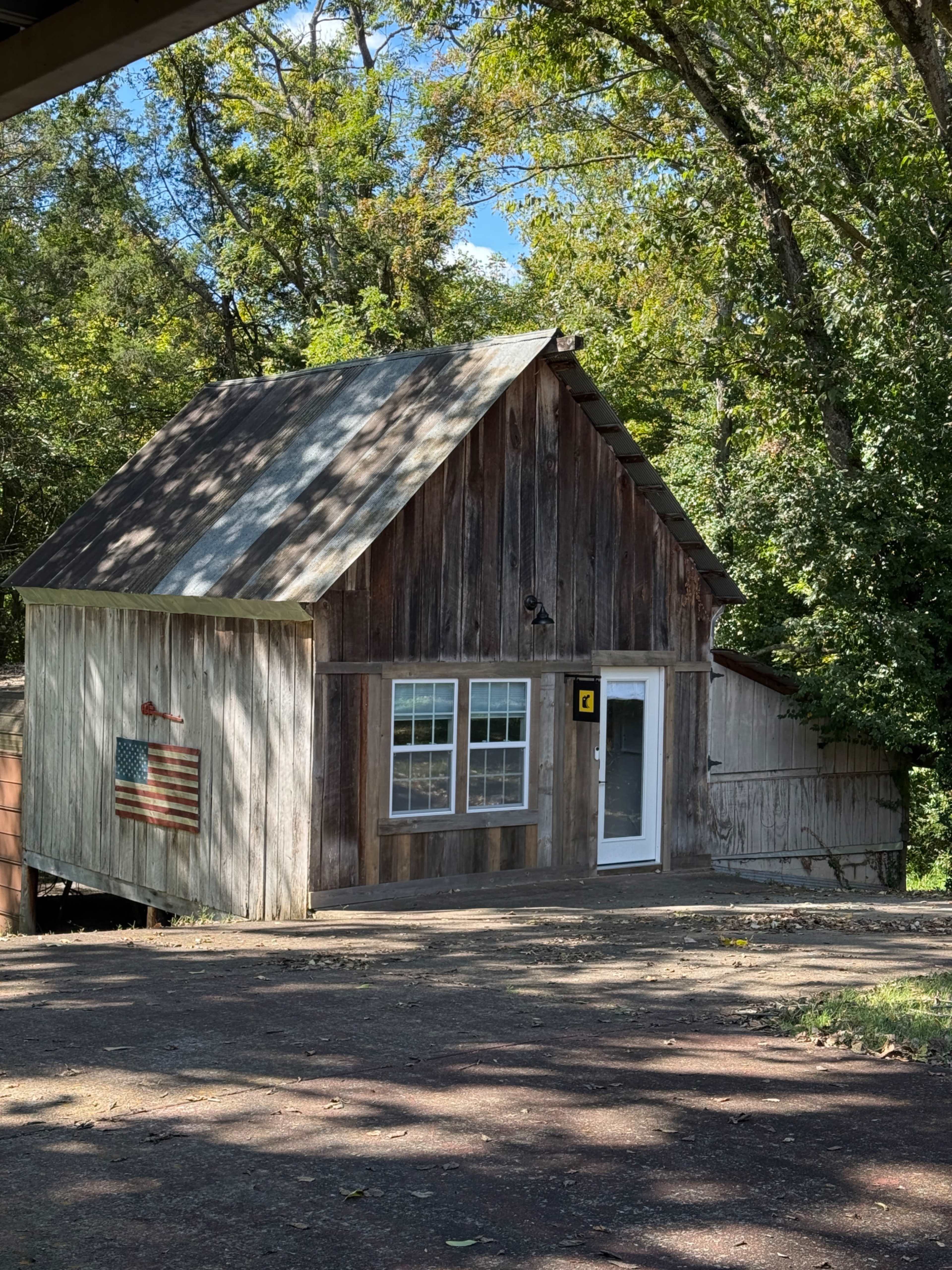 The image shows a rustic wooden cabin with a front porch, large windows, and a small American flag attached to its side, set amidst trees under a clear blue sky.